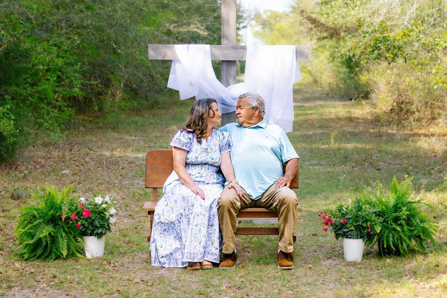 Older couple sitting on a bench in front of a wooden cross surrounded by potted plants and flowers.