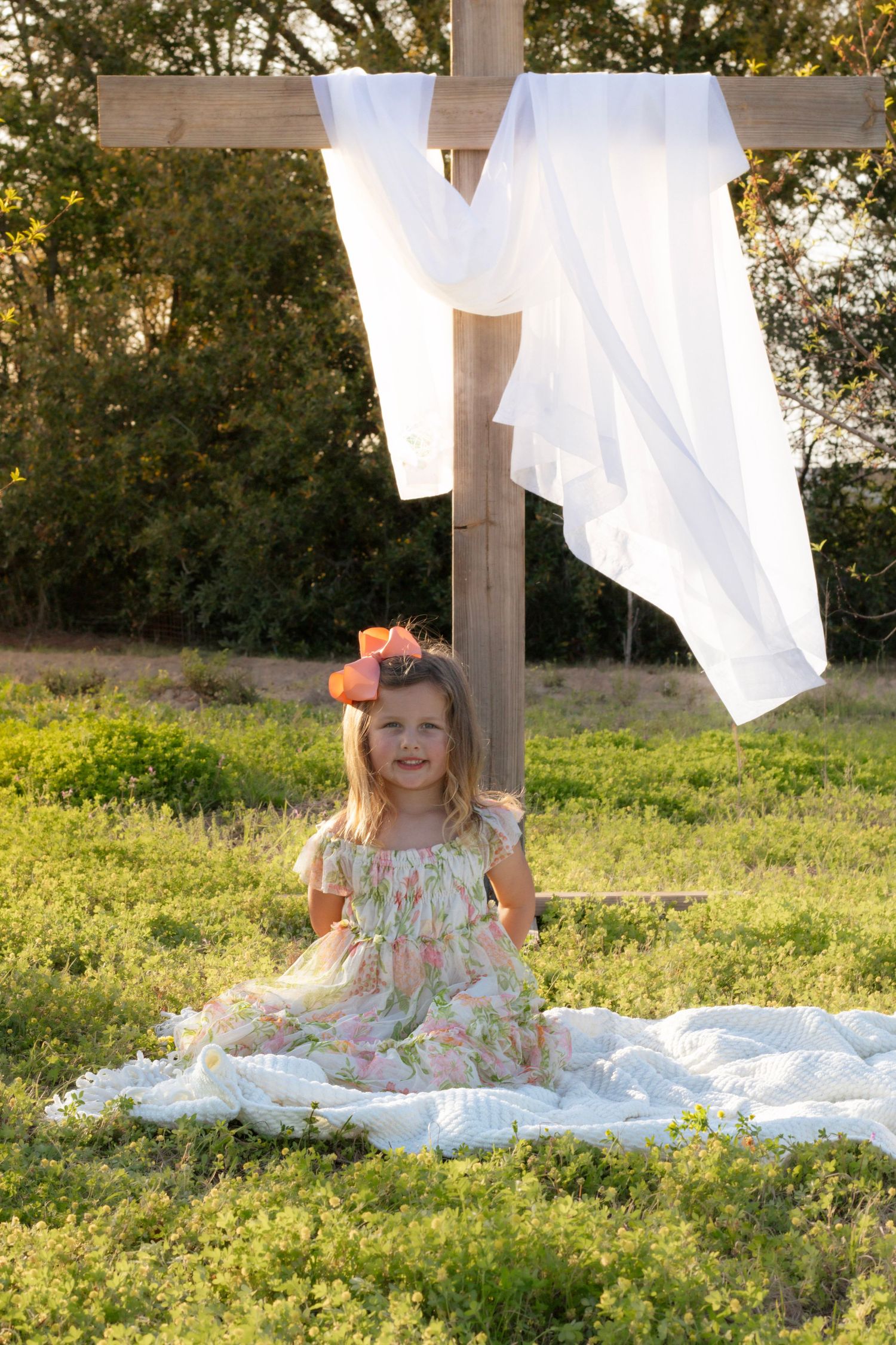 Smiling little girl in floral dress with pink bow sitting beside a wooden cross in a meadow.