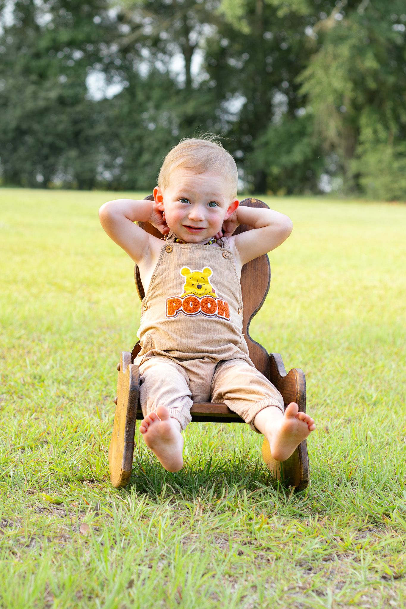 Smiling toddler in Winnie the Pooh outfit relaxing in wooden chair on green grass outdoors.