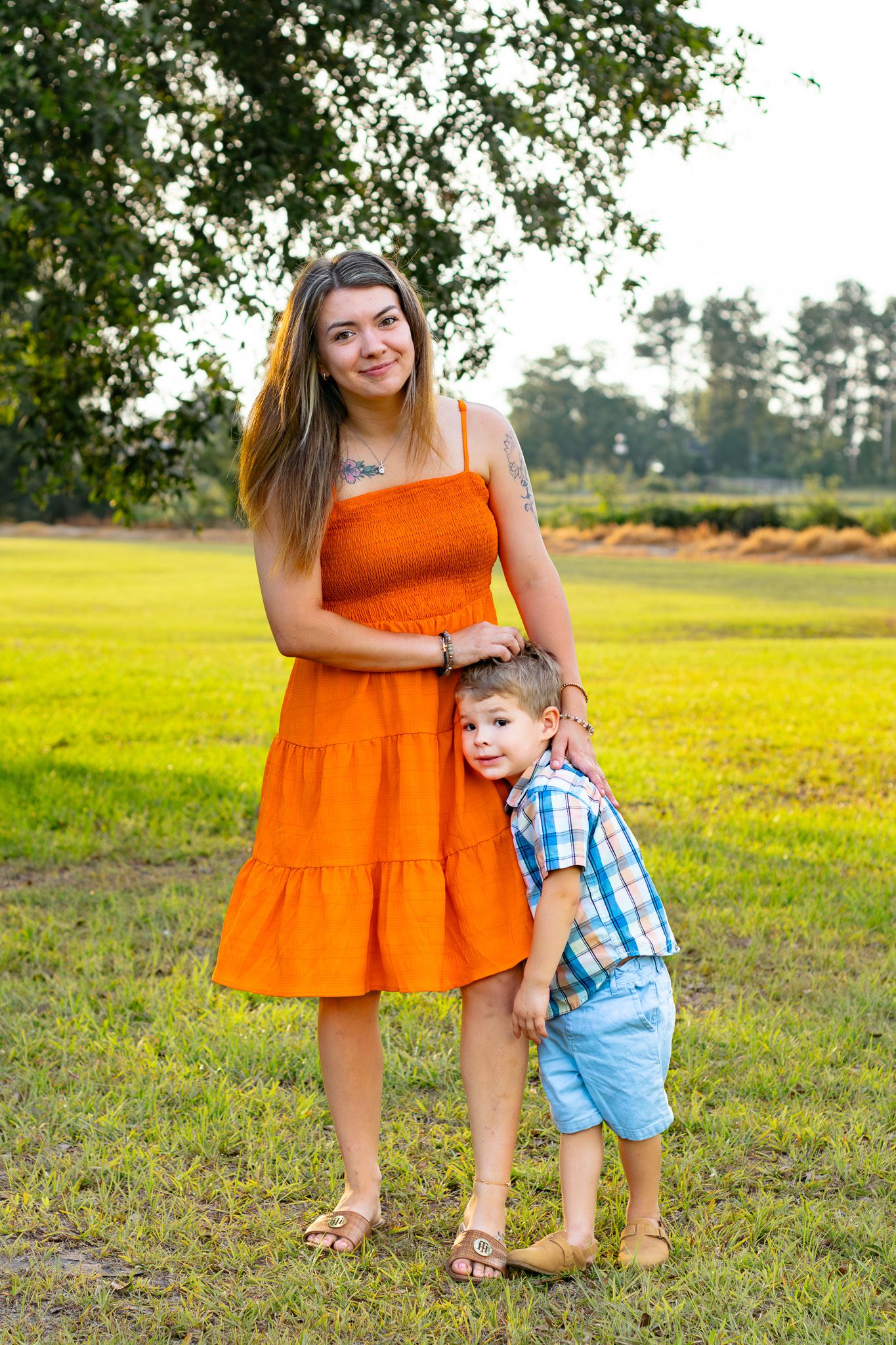 A young woman in an orange dress stands with a toddler boy in a sunny green park setting.