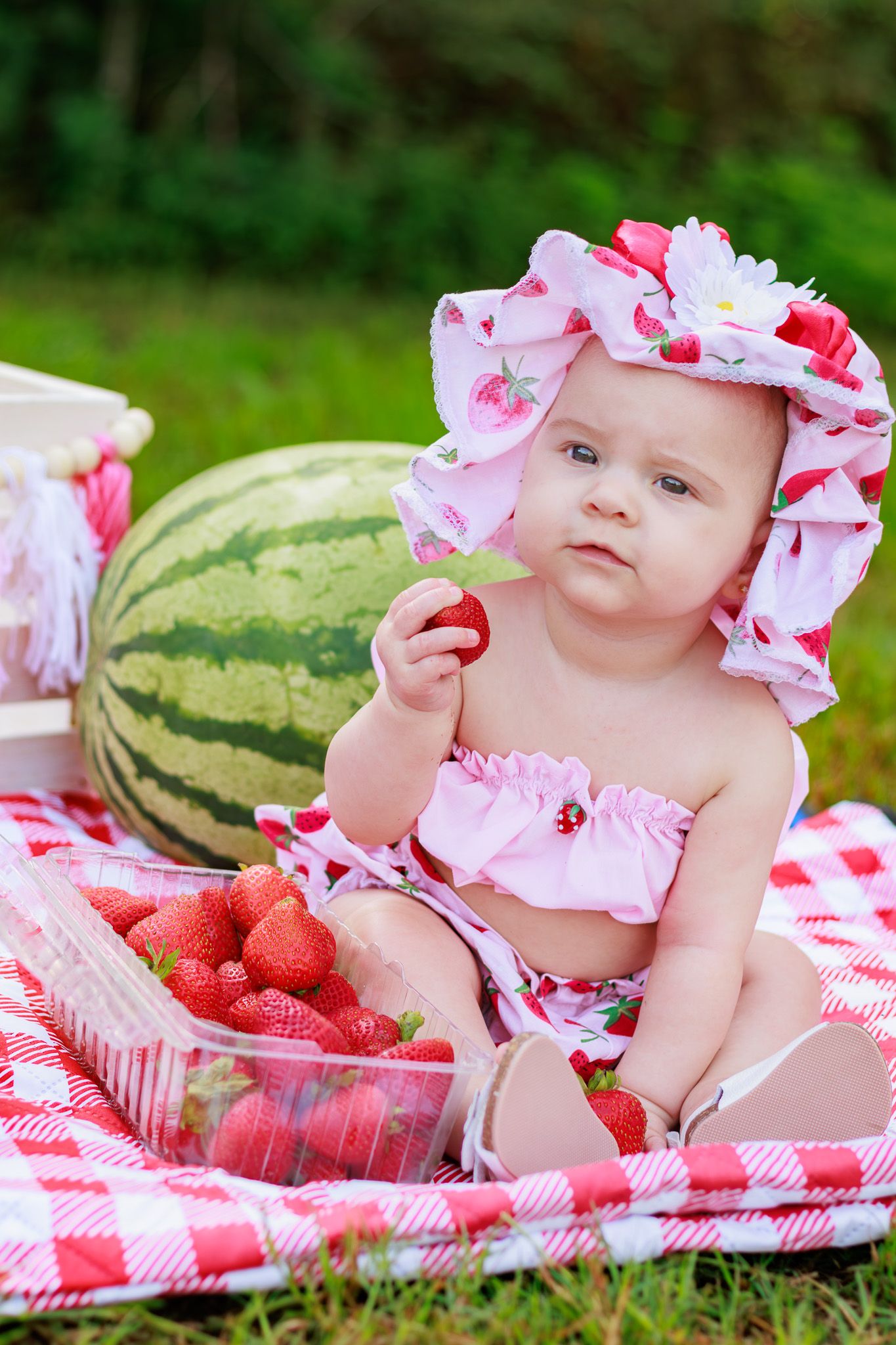 Baby girl in strawberry hat sits on picnic blanket holding strawberry with watermelon and fresh strawberries nearby.