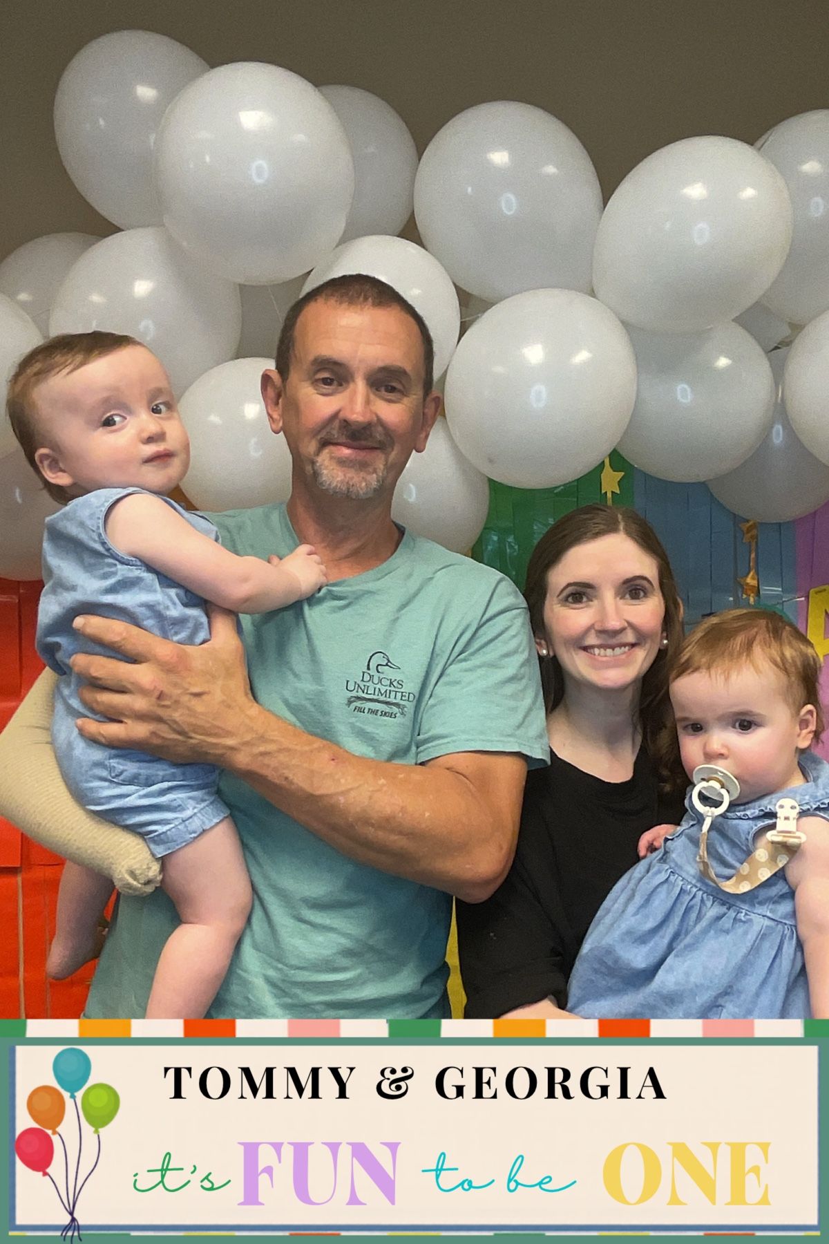 Parents holding twin babies at a colorful first birthday party with white balloon backdrop.