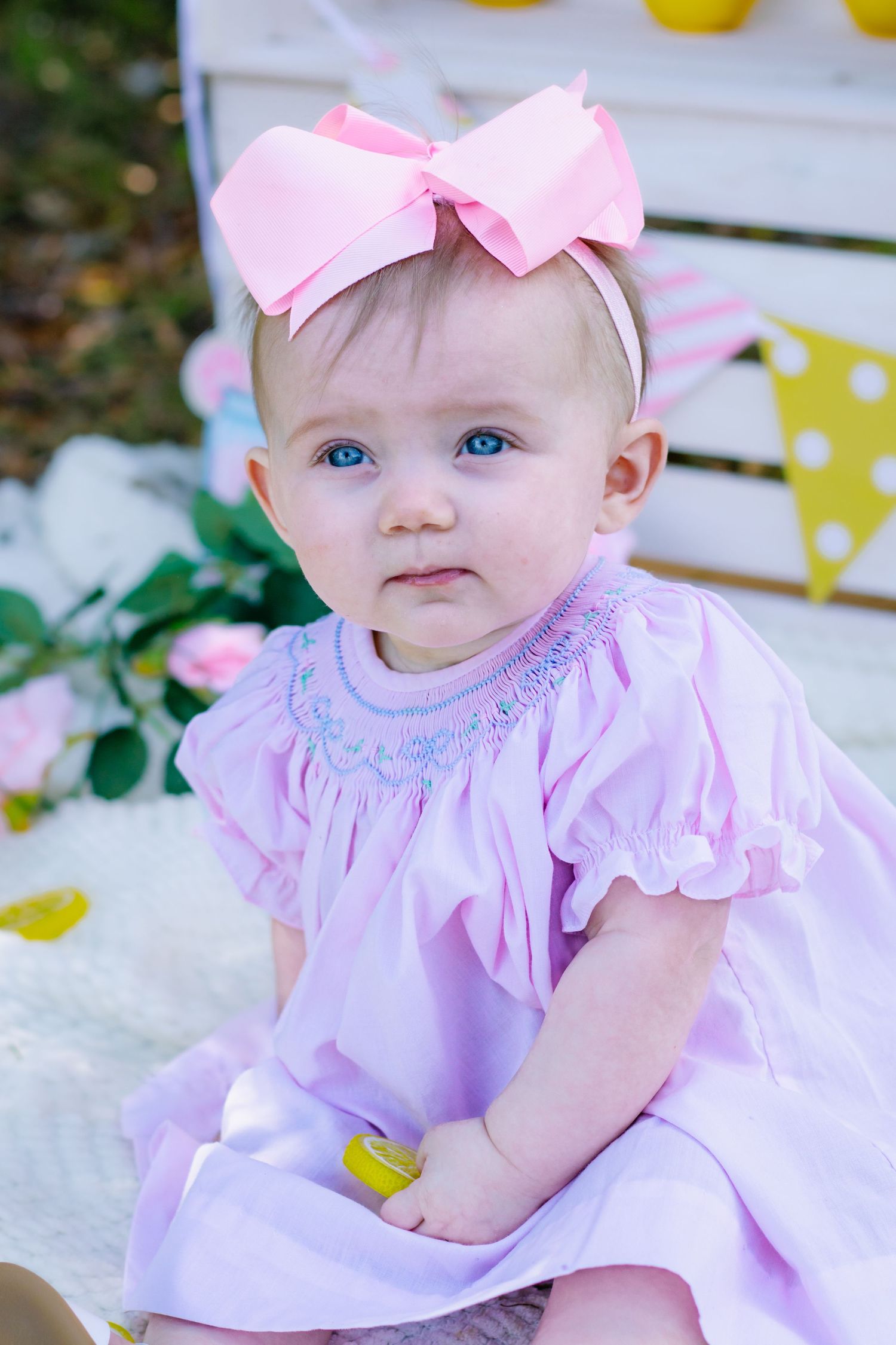 Blue-eyed baby girl in pink smocked dress with large bow headband sitting at outdoor birthday setup.