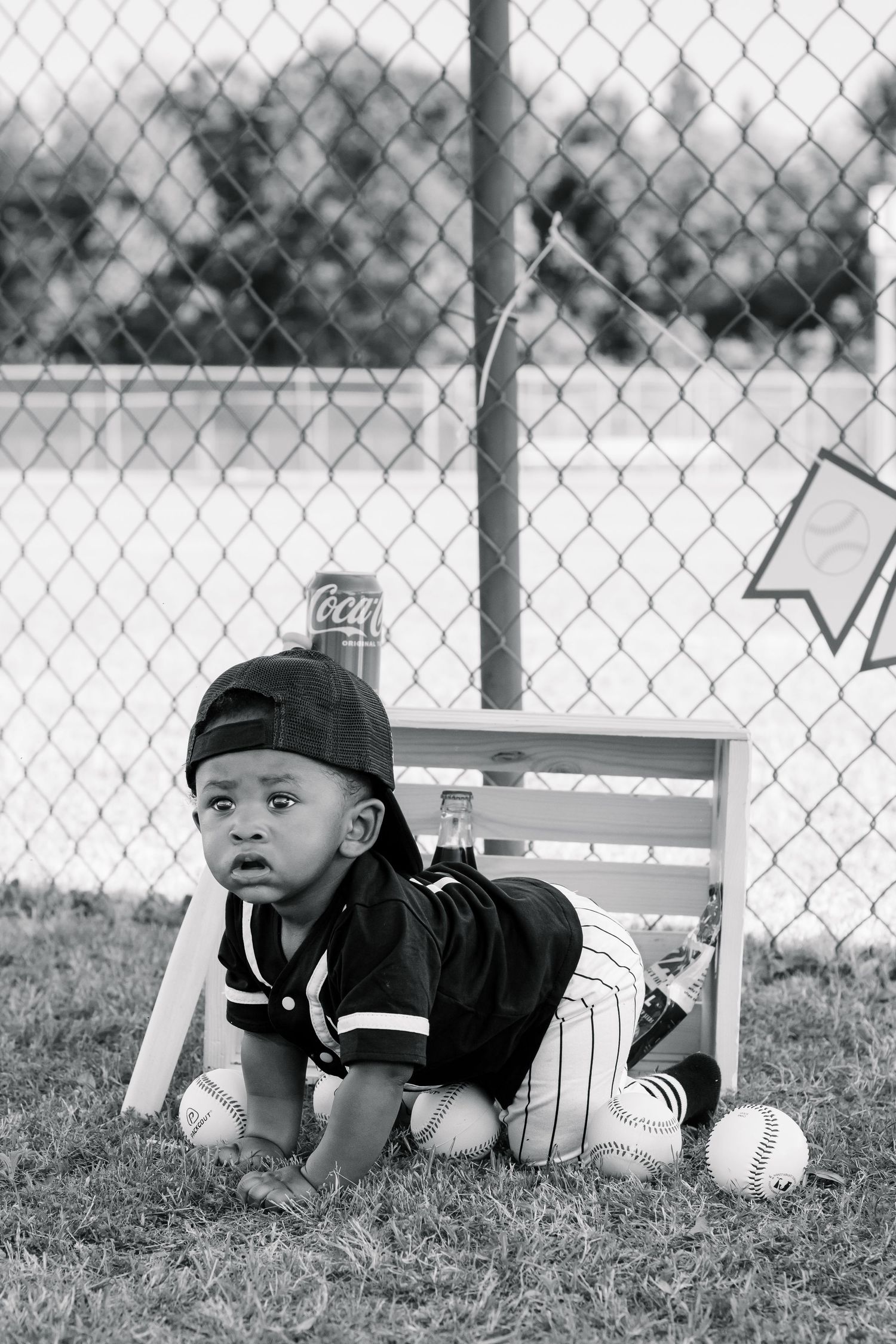 Black and white photo of a baby in baseball uniform crawling on grass with baseballs near a chain-link fence.