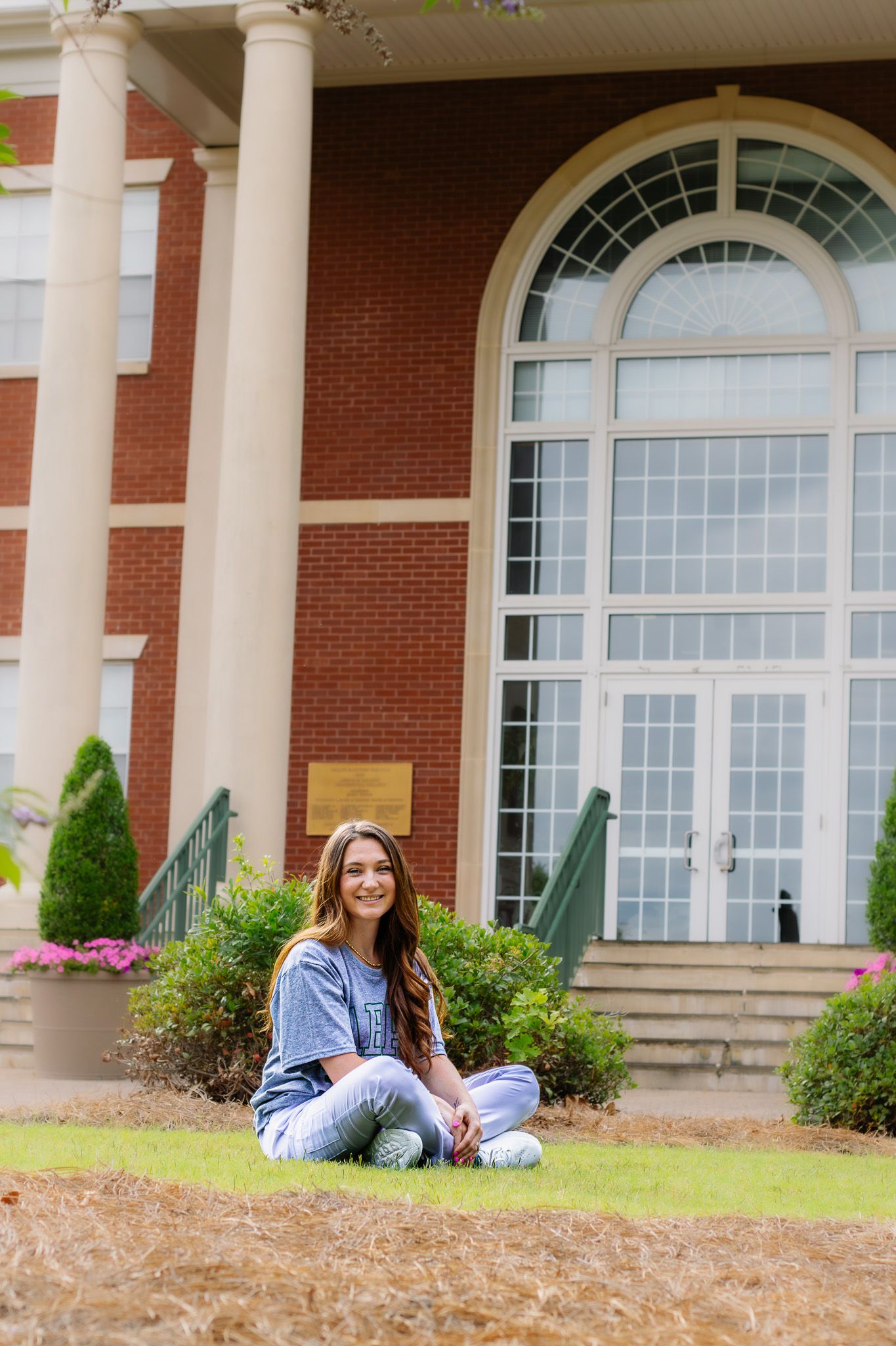 Young woman sits cross-legged on grass in front of red brick college building.