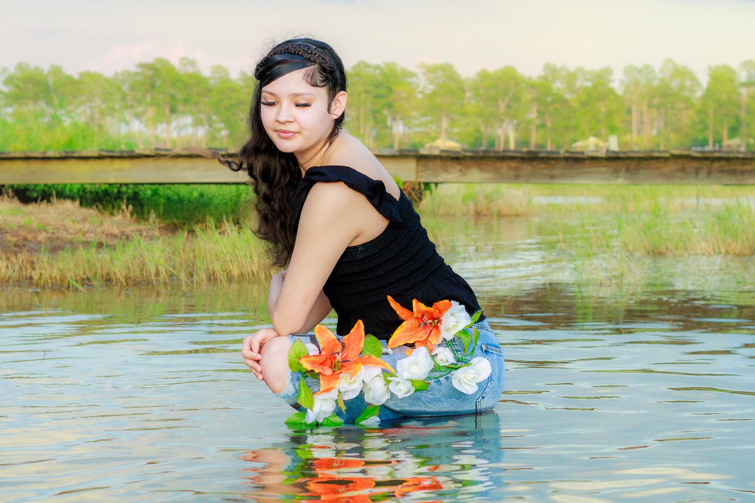 Young woman in black top and floral skirt kneeling in calm water near a grassy riverbank.