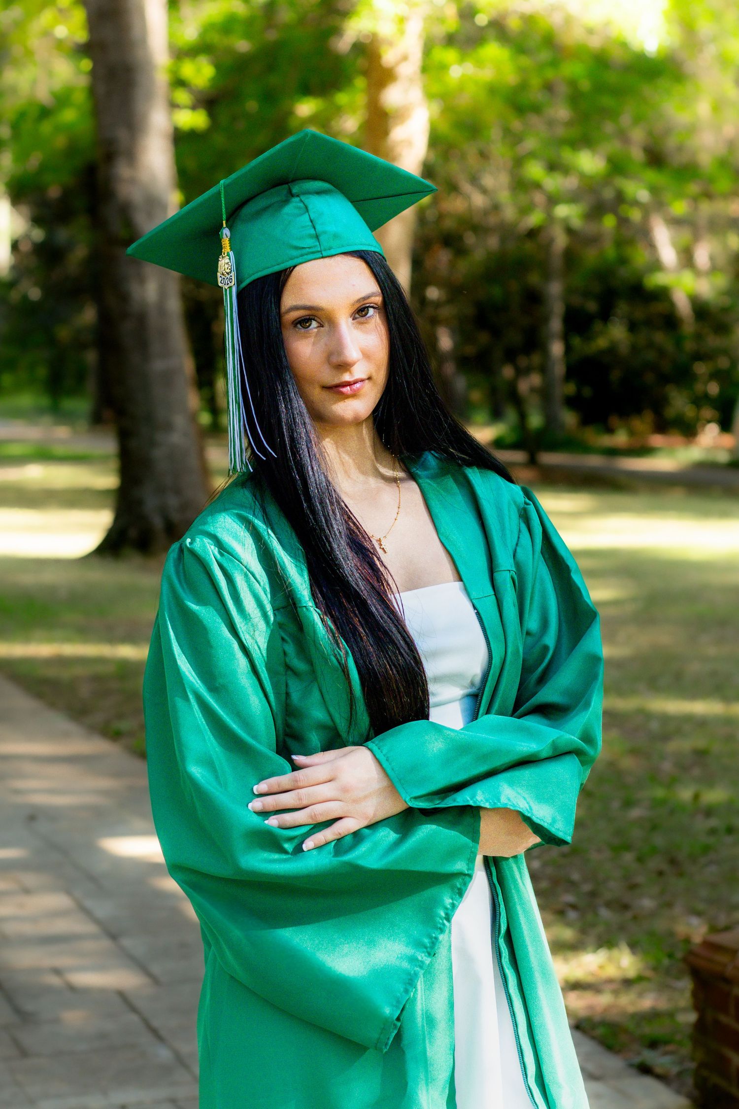 Young woman in green graduation cap and gown stands with arms crossed in sunny outdoor park setting.