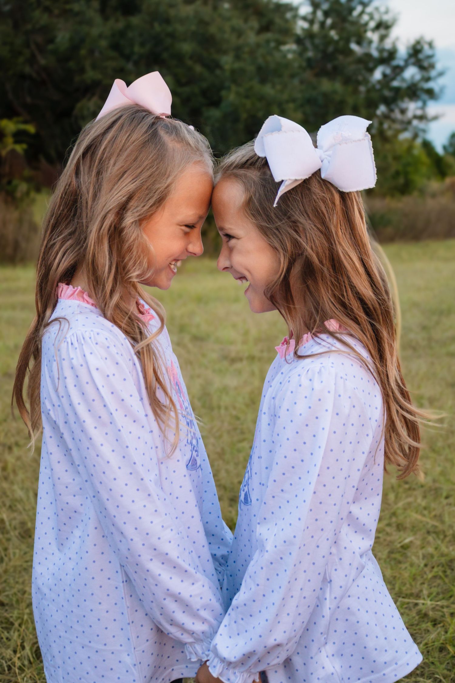 Two smiling girls in matching polka dot dresses touching foreheads playfully outdoors.