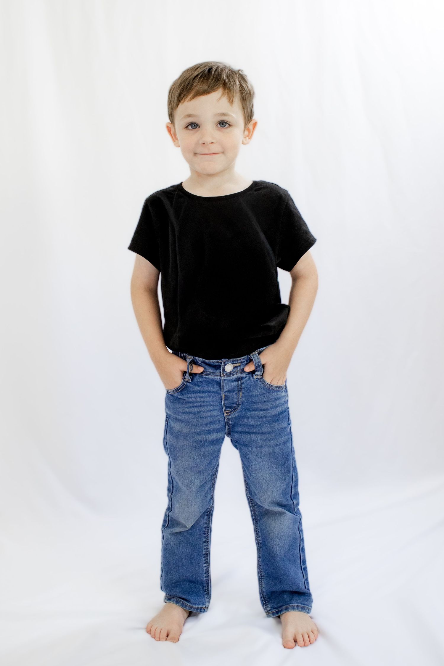 Young boy in a black t-shirt and blue jeans standing with hands in pockets, smiling against a white background.