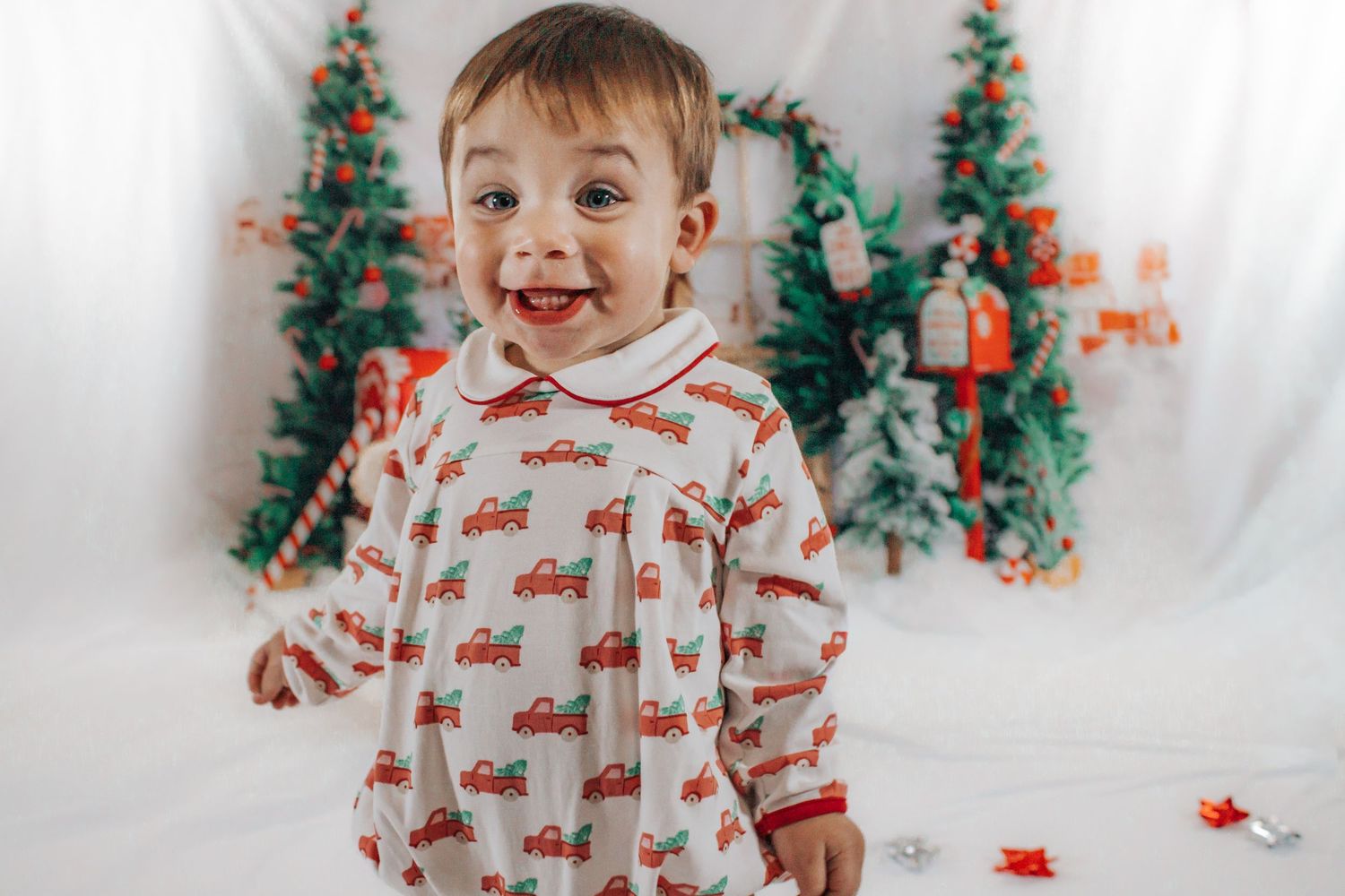 Happy toddler boy in Christmas pajamas smiles in front of festive holiday backdrop.