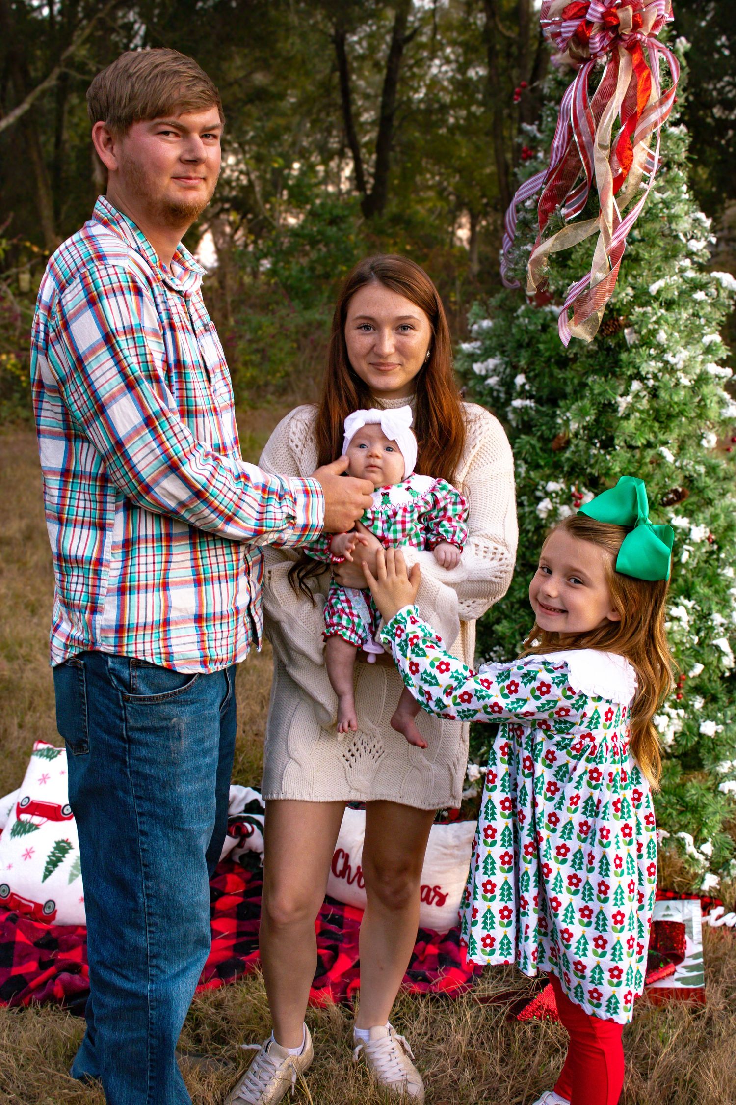 Family Christmas photo with parents holding newborn baby and young girl in holiday outfits beside decorated tree.