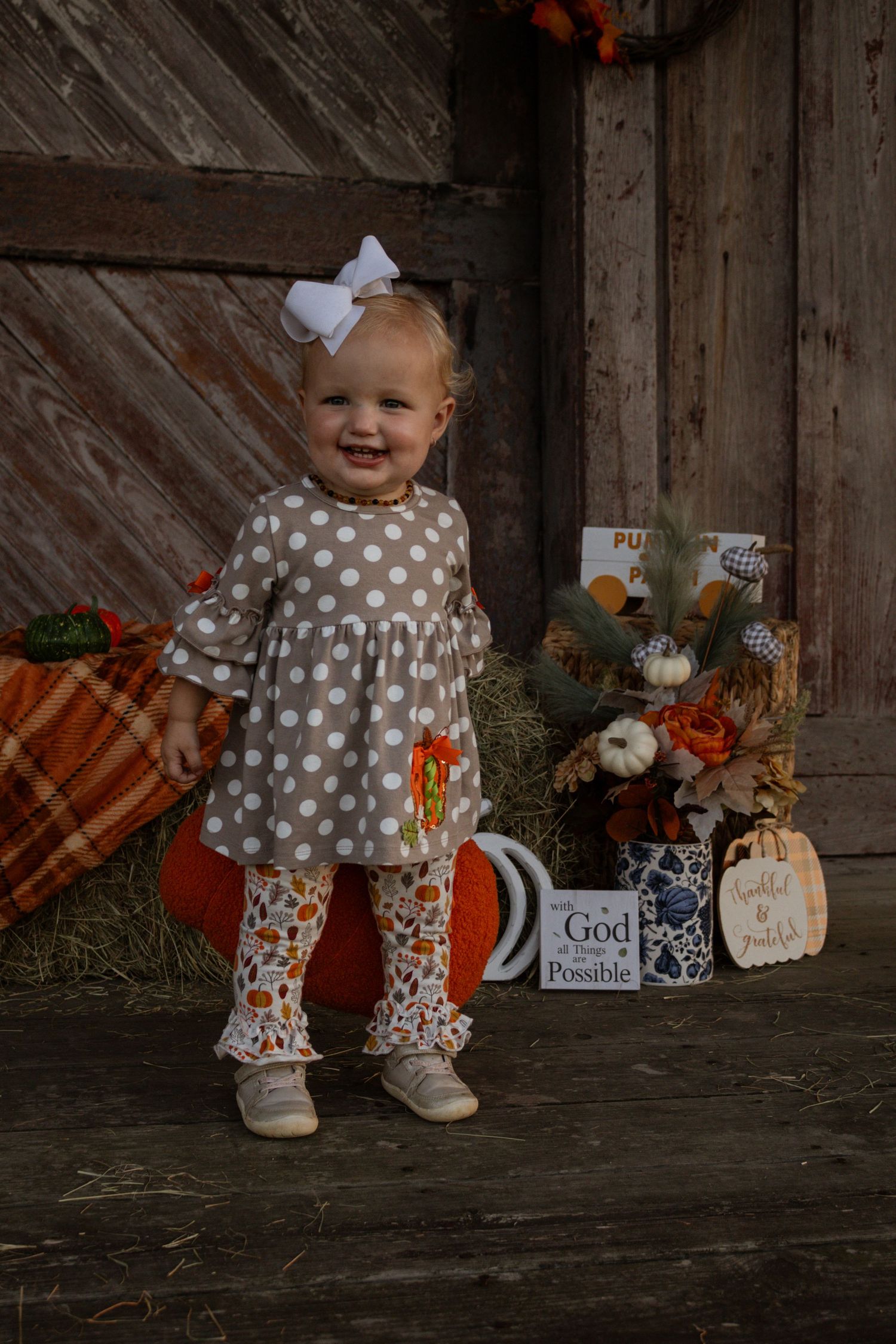 Happy toddler girl in polka dot outfit standing by pumpkins in rustic fall barn photo.