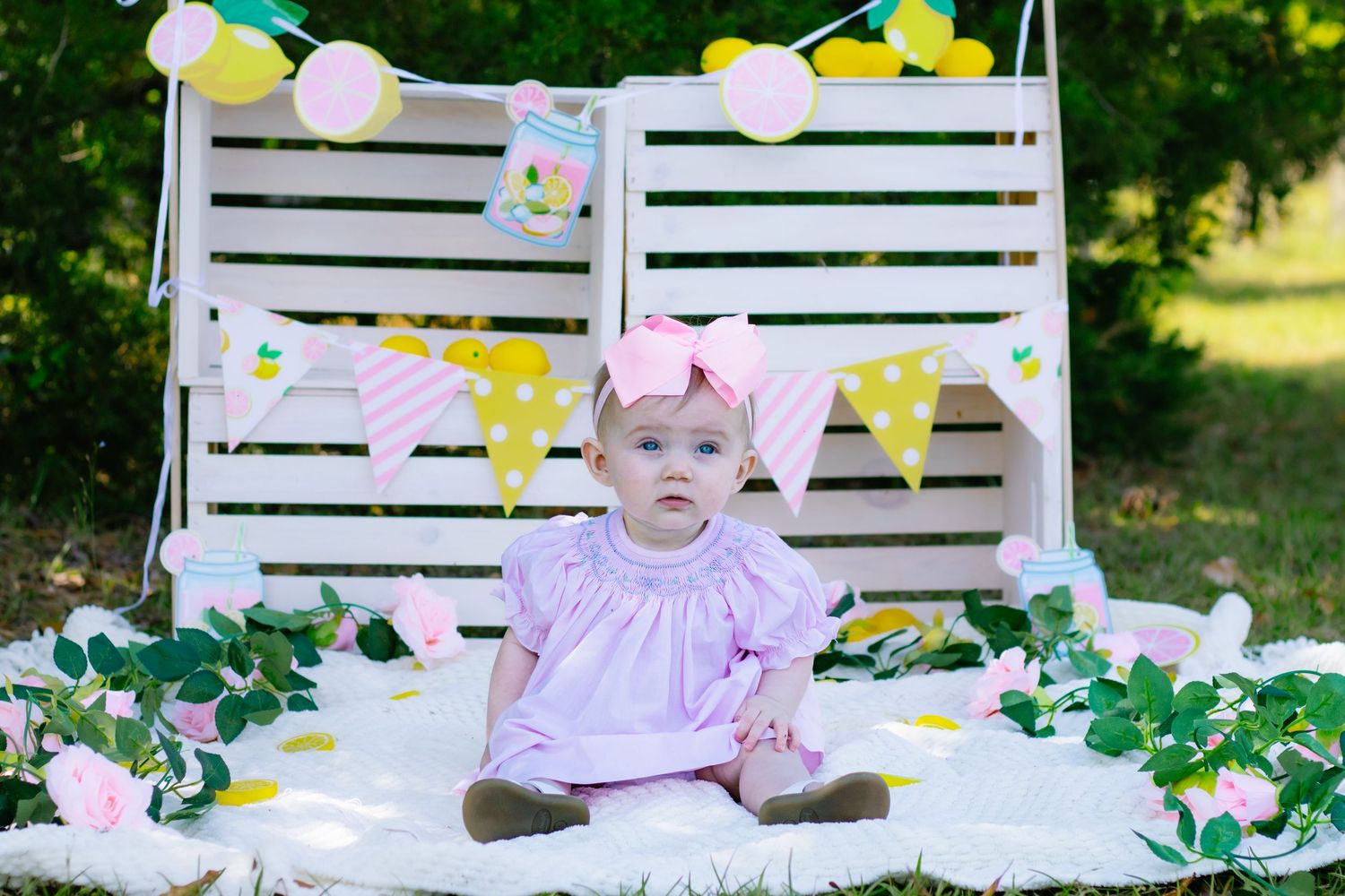 Baby girl in pink dress with bow headband sitting at outdoor lemon-themed first birthday setup.