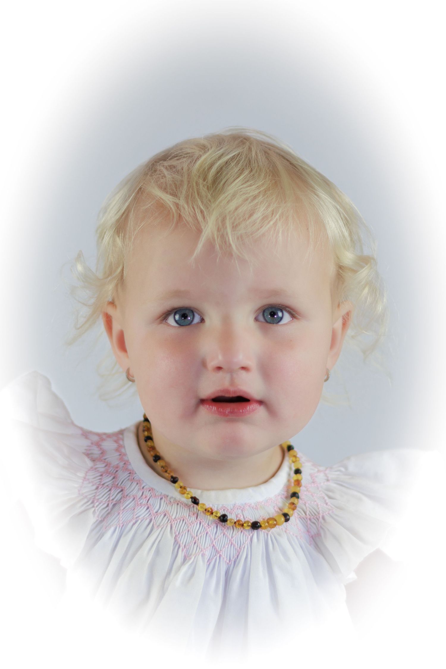 Blonde toddler girl with blue eyes wearing a white smocked dress and amber beaded necklace in a portrait photo.