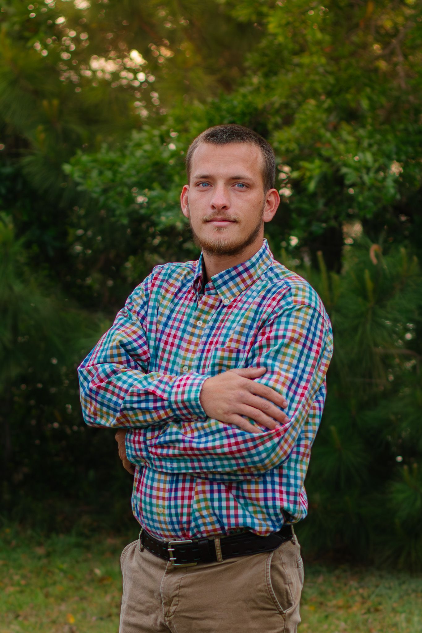 Young man with crossed arms wearing colorful plaid shirt standing outdoors with green trees in background.