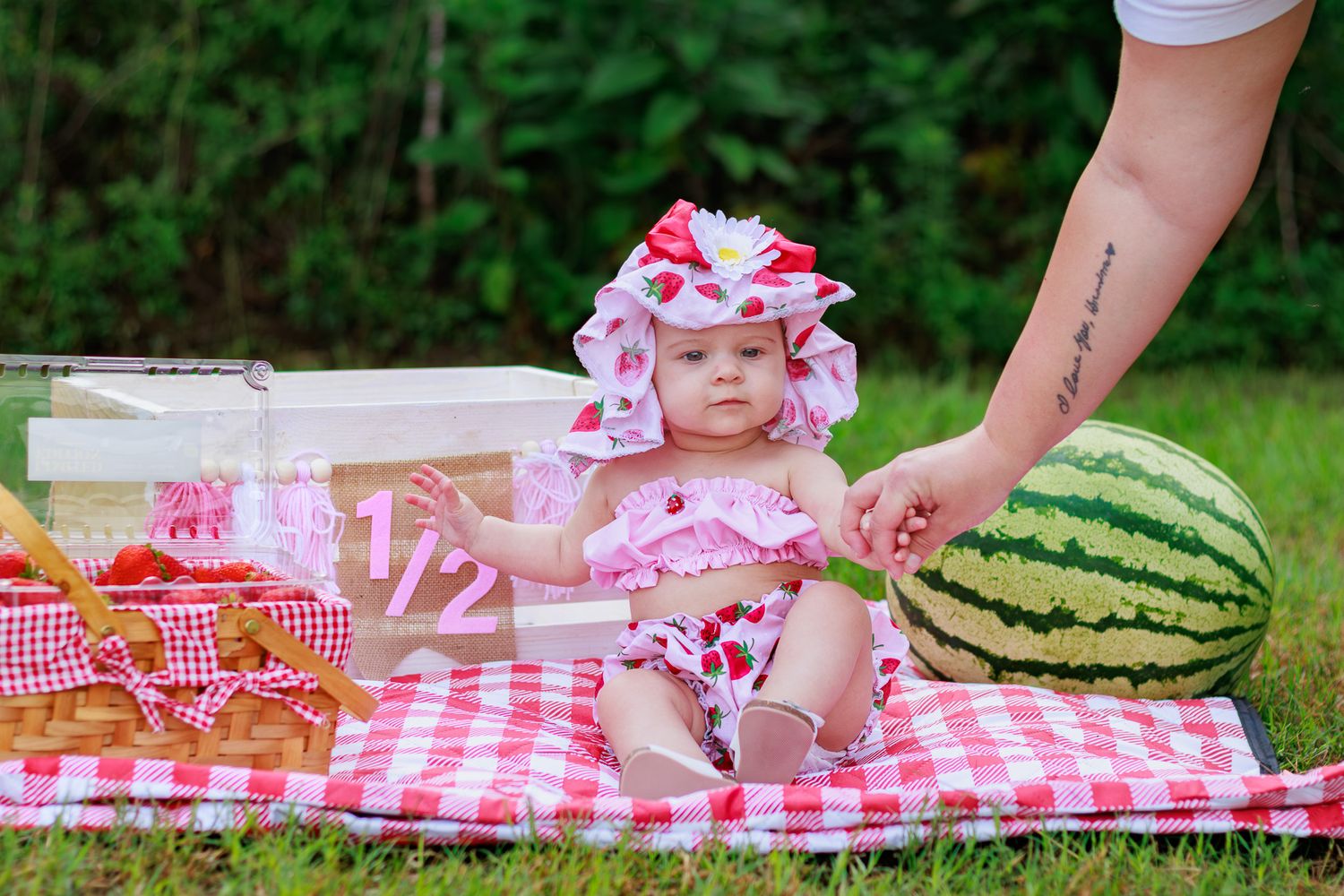 Adorable baby girl in strawberry outfit sits on gingham blanket beside watermelon for half birthday photo.