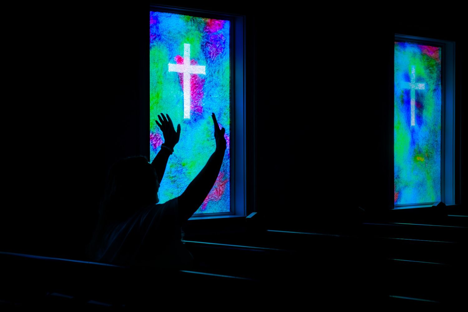 Silhouette of person raising hands in worship before glowing stained glass cross windows.