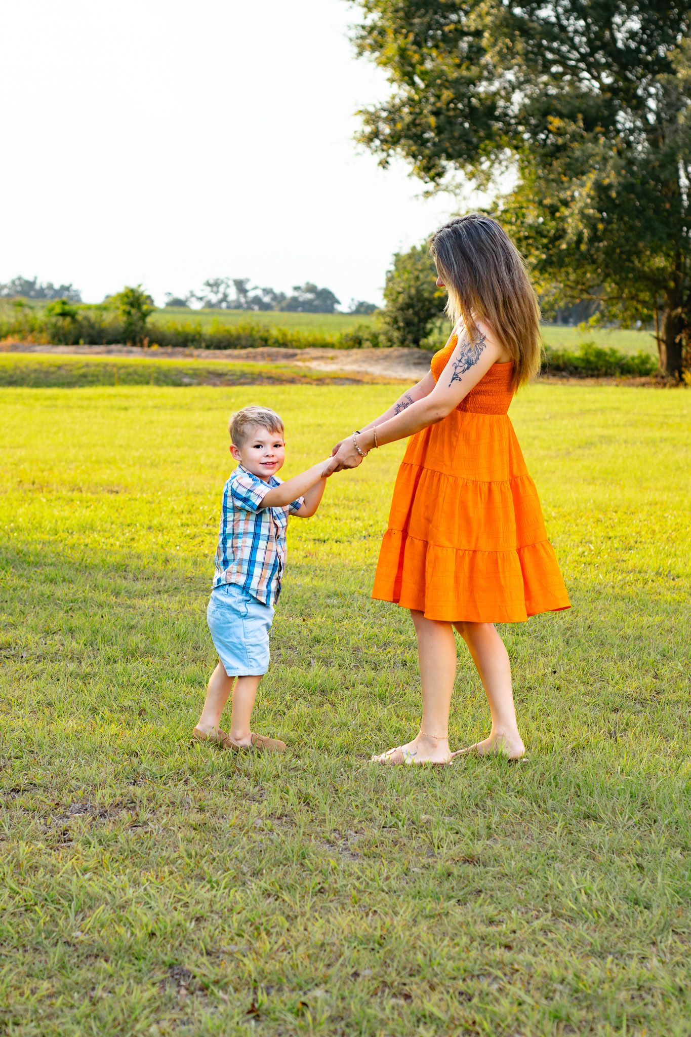 A woman in an orange dress holds hands with a young boy in a plaid shirt while playing in a green field.