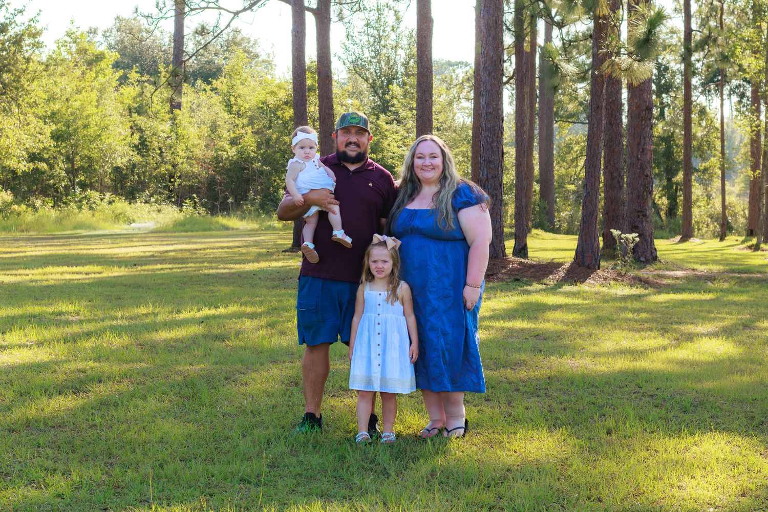 Family of four poses outdoors on green grass surrounded by tall pine trees on a sunny day.