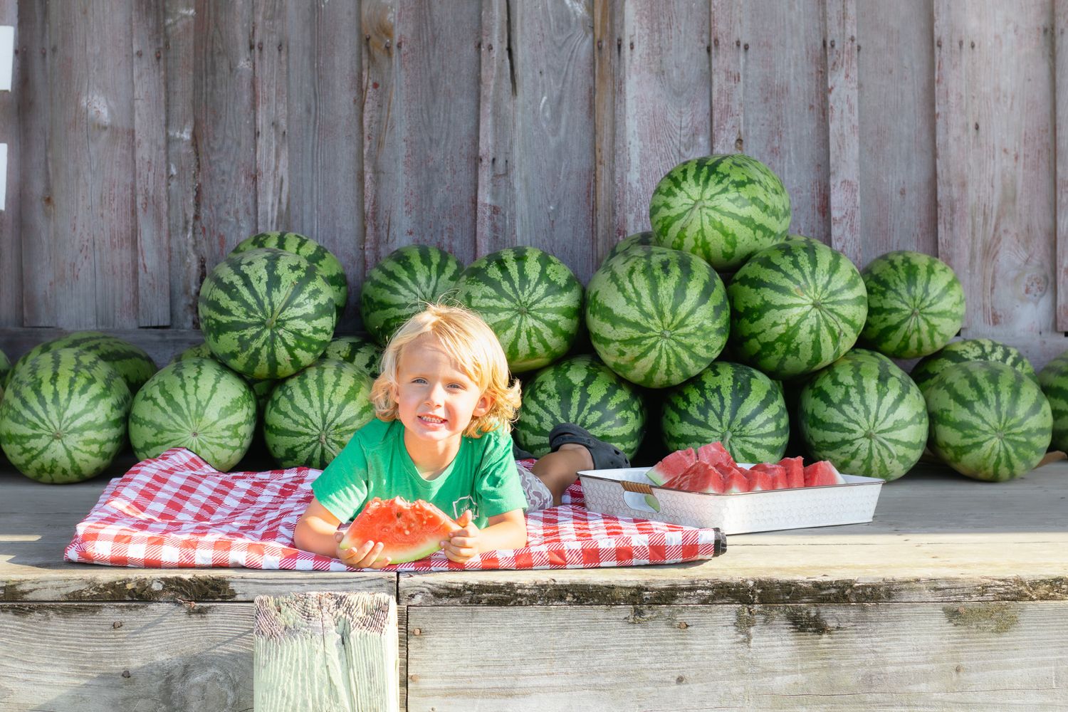 A cheerful blond child smiles at a picnic table surrounded by a large pile of fresh watermelons outdoors.