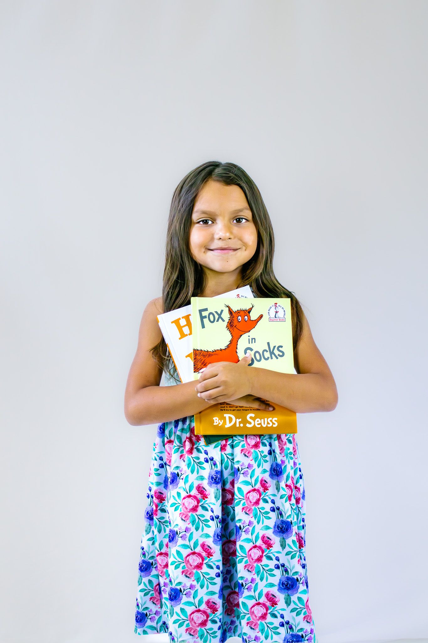 Young girl in floral dress smiling while holding a Dr. Seuss Fox in Socks book against a gray background.