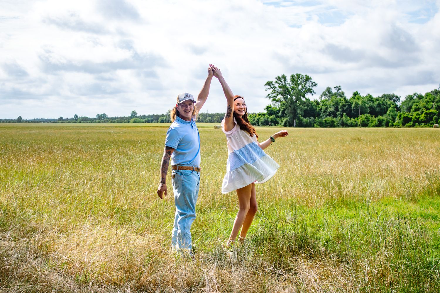 Joyful couple dancing and spinning in a golden grassy field with trees and cloudy sky in the background.