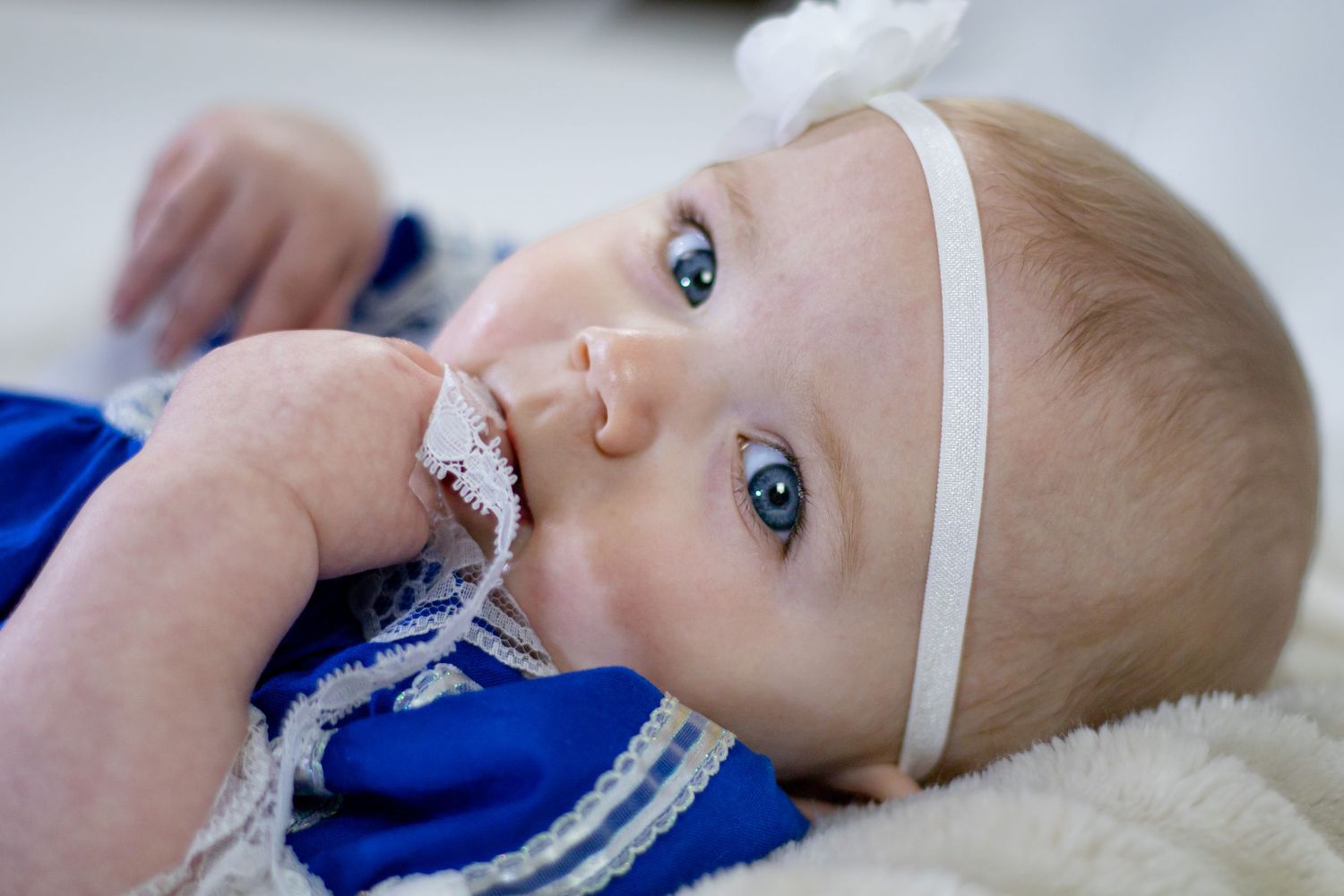 Sweet blue-eyed baby girl in blue dress with white flower headband lying down chewing lace.
