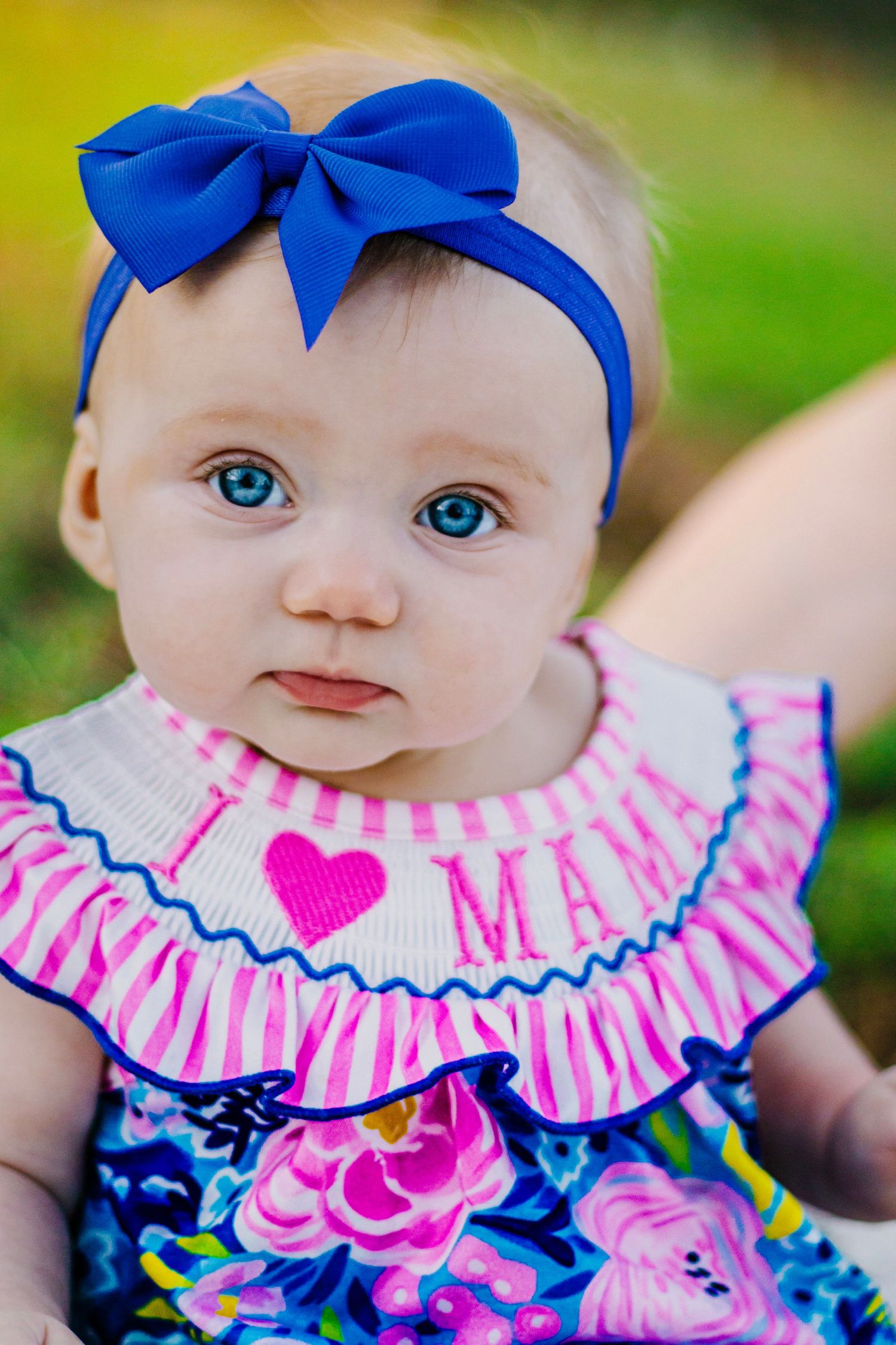 Close-up portrait of blue-eyed baby girl wearing blue bow headband and floral I Love Mama outfit.