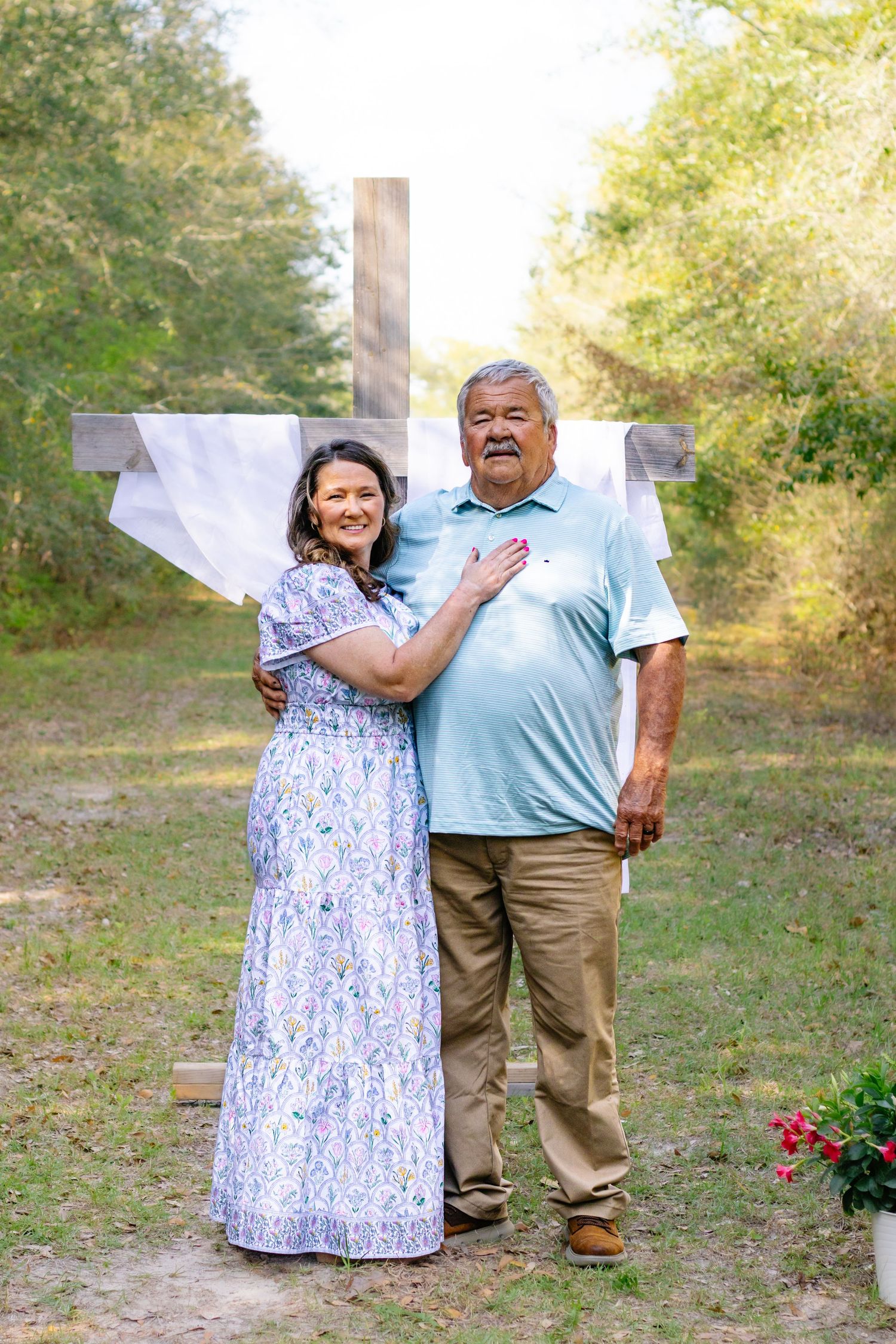 Couple posing in front of a wooden cross outdoors, woman in floral dress embracing man in blue polo.