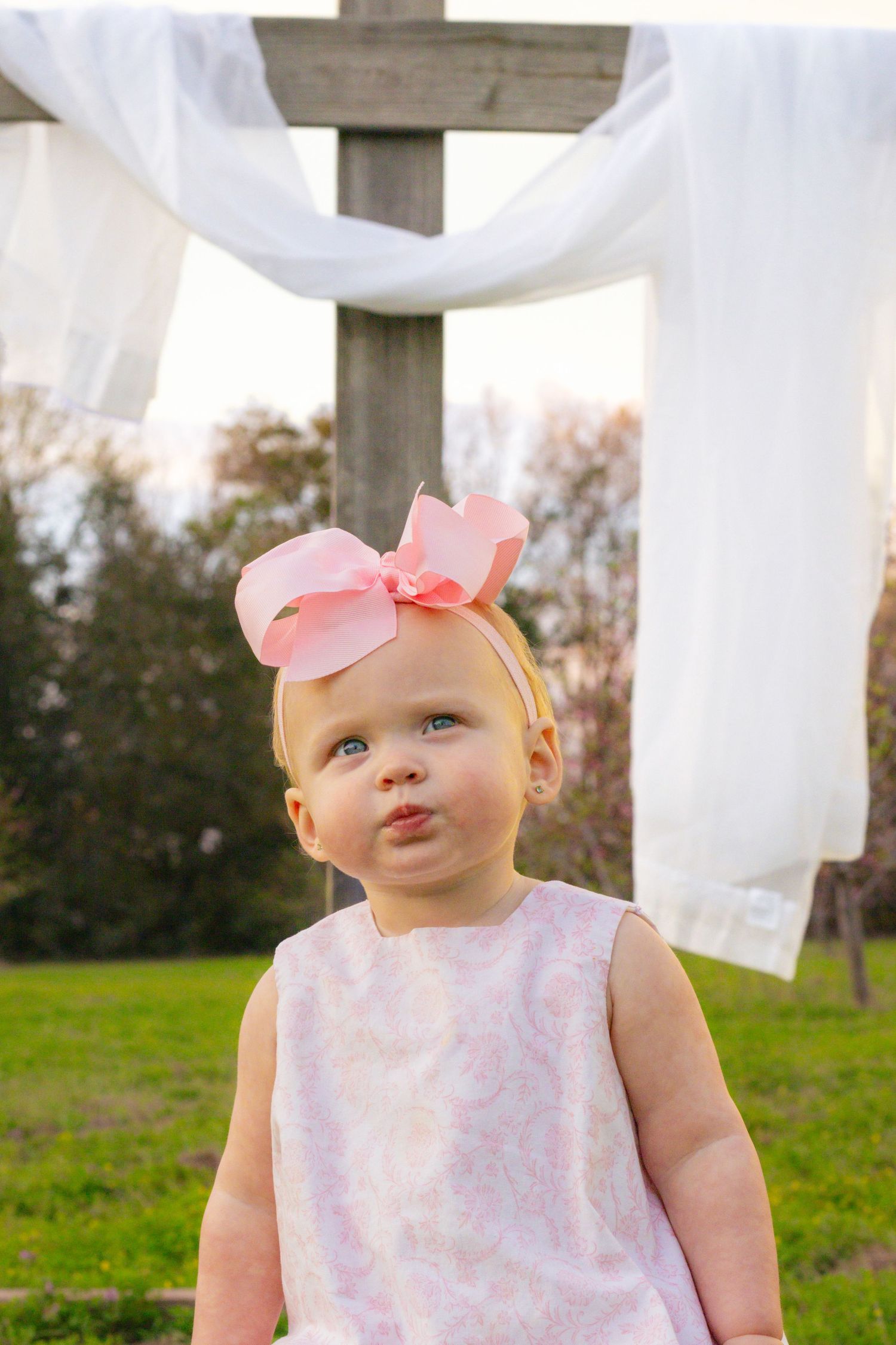 Adorable baby girl in pink bow headband and white dress poses outdoors near a wooden cross with white fabric.