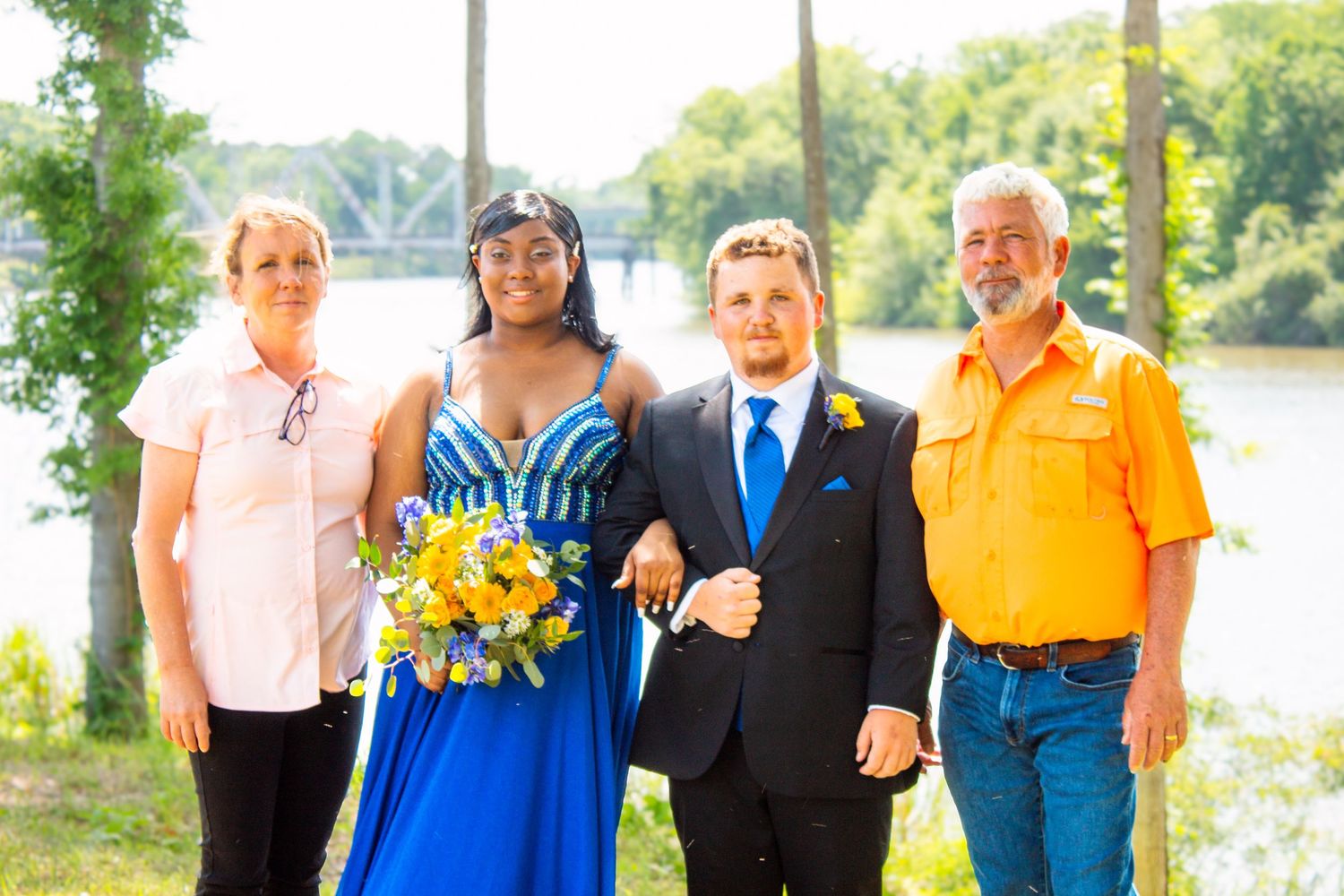 Family of four posing outdoors near river, young couple in prom attire with older parents.