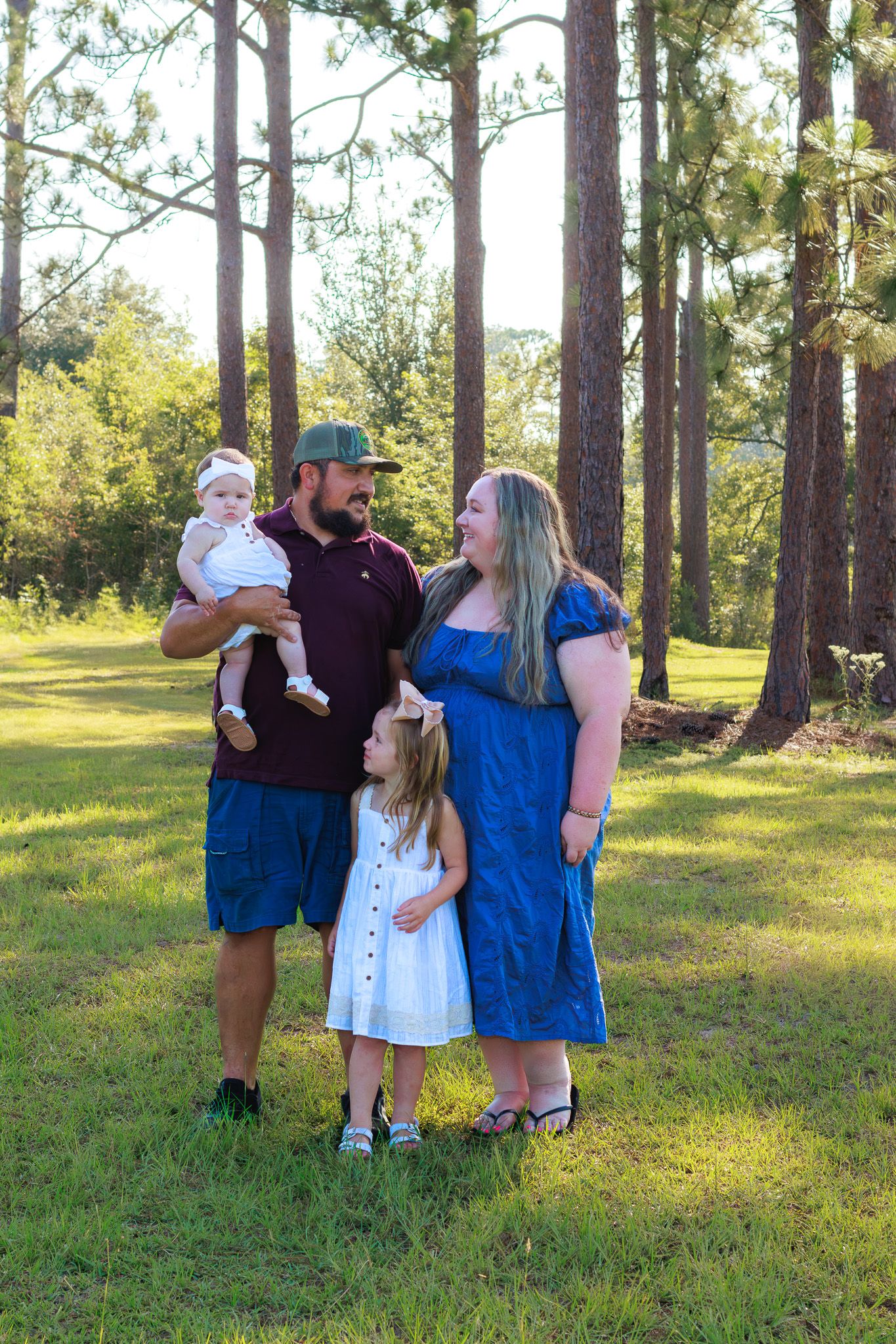Family of four posing outdoors among tall pine trees on green grass in sunny weather.
