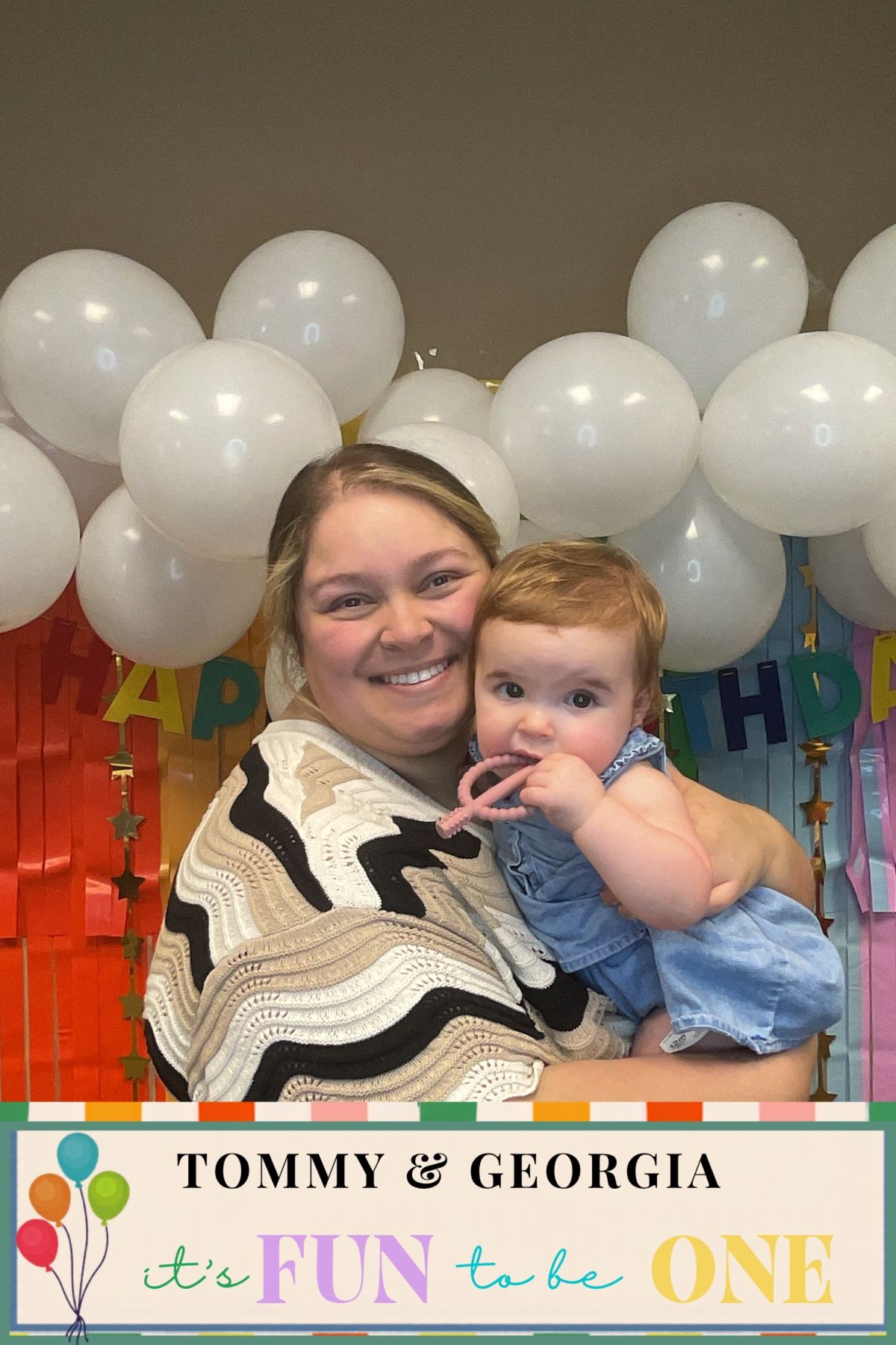Woman holding baby girl at Tommy & Georgia's first birthday party with white balloon backdrop.