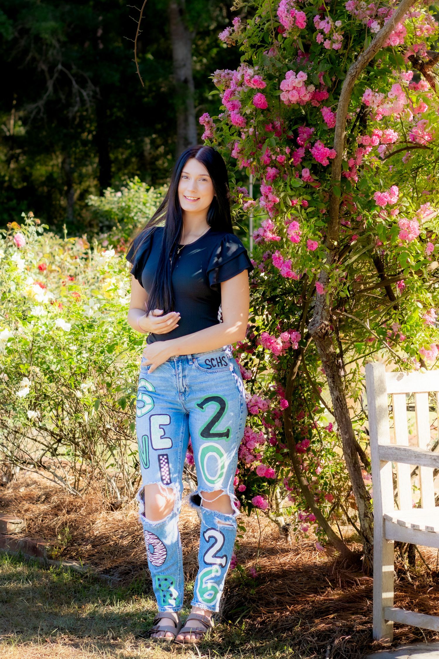 Young woman in decorated jeans and black top poses near pink flowering roses beside a white bench outdoors.