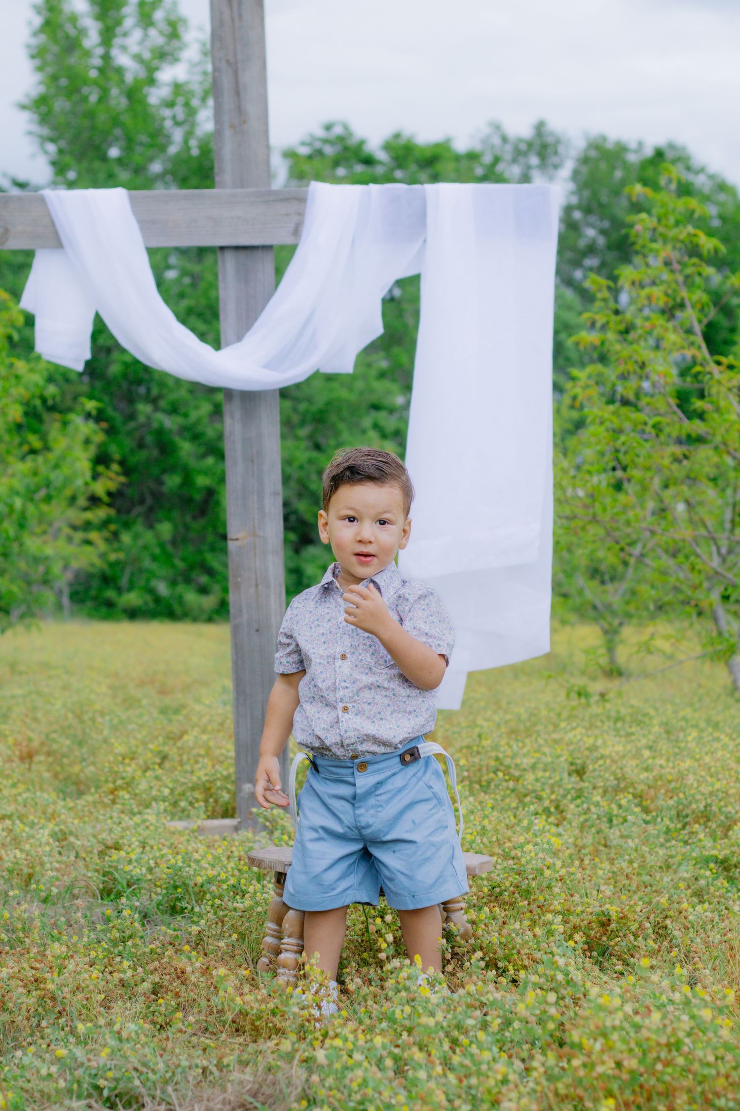 Young boy in floral shirt and denim shorts standing outdoors near wooden cross with white fabric draped over it.