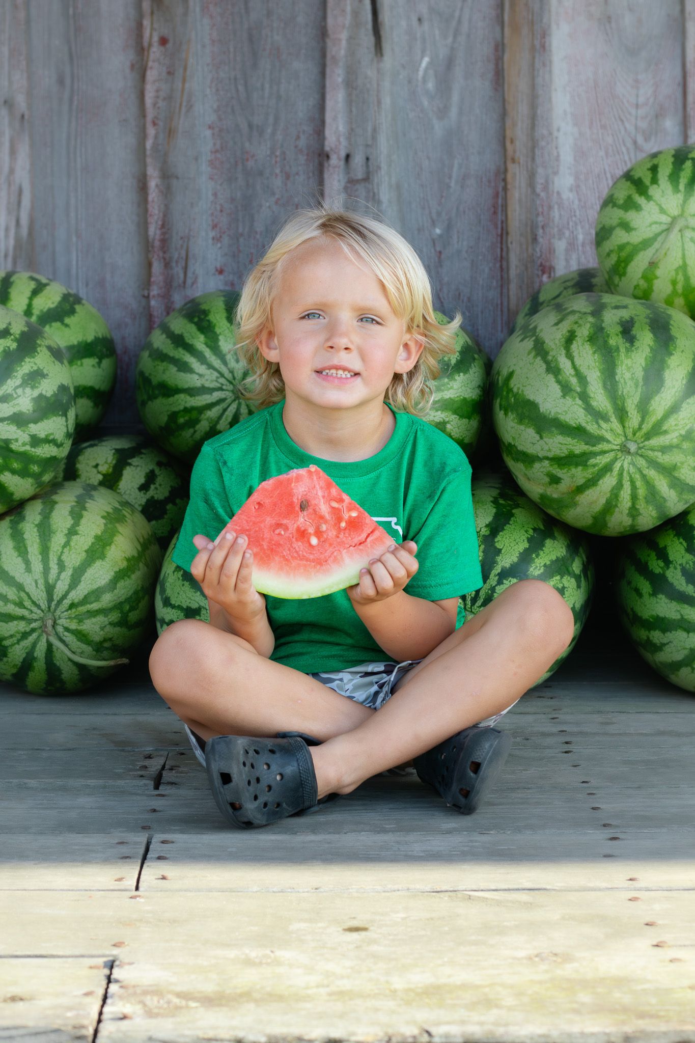 Smiling blond boy sitting cross-legged holding a watermelon slice, surrounded by whole watermelons against a wooden wall.