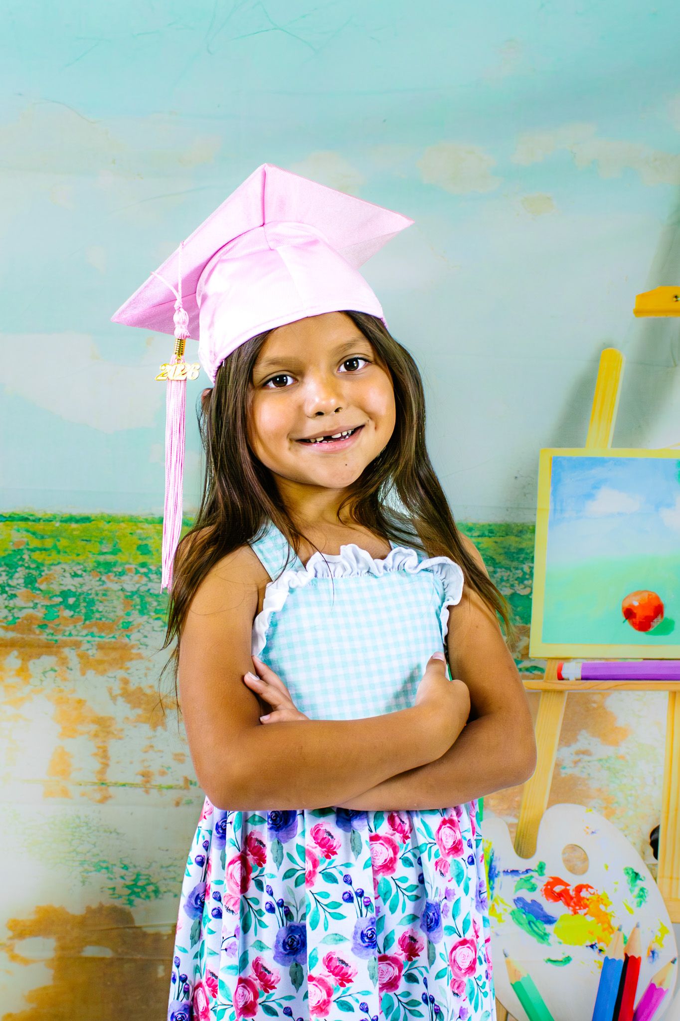 A smiling young girl wearing a pink graduation cap and floral dress holds a book while standing near an easel.