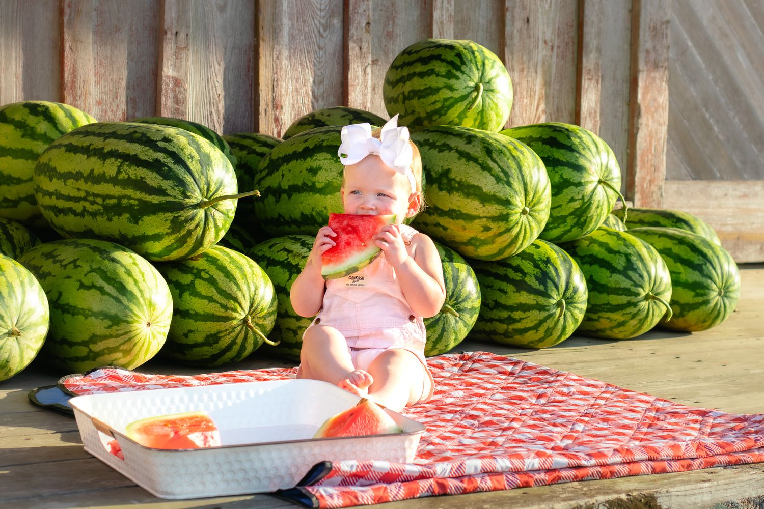 Smiling baby girl in pink romper enjoying fresh watermelon slice in front of stacked watermelons.