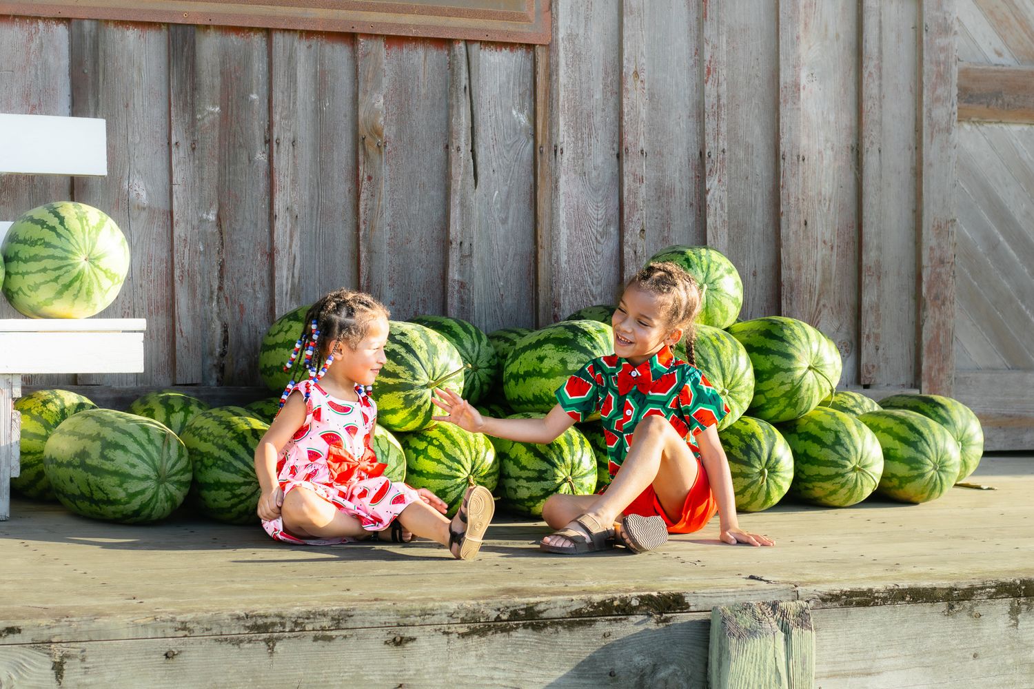 Two young girls in colorful dresses sitting together beside a large pile of watermelons outdoors.