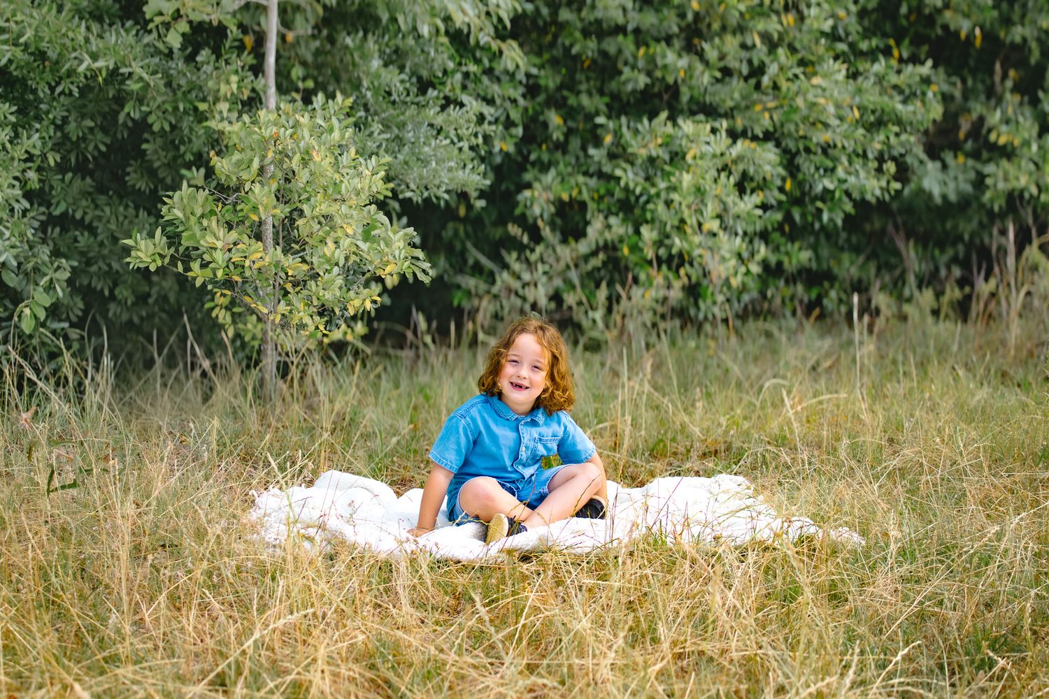 Happy curly-haired child sitting on a white blanket in a golden grassy field surrounded by trees.