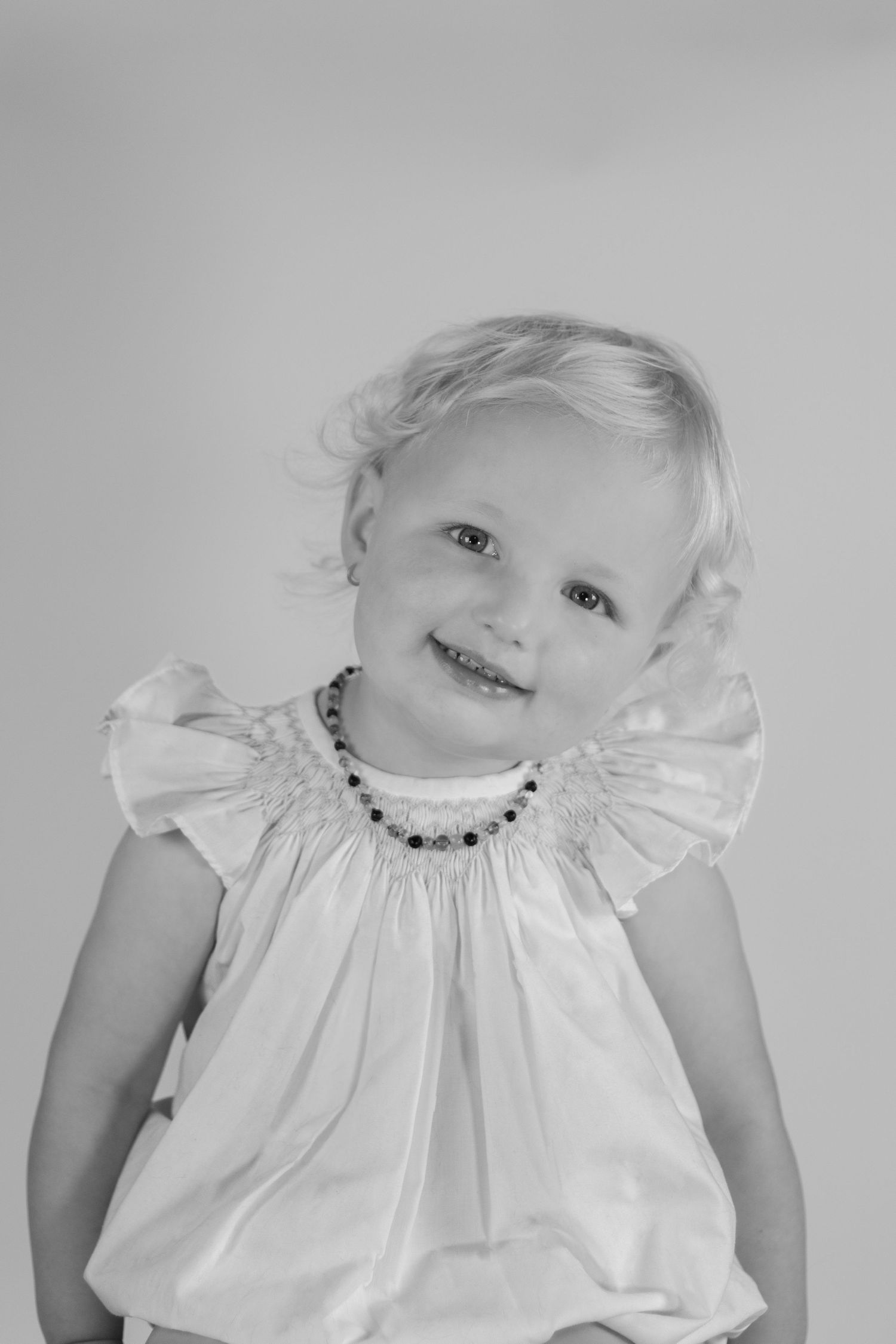 Black and white portrait of a smiling toddler girl wearing a white ruffled dress and beaded necklace.