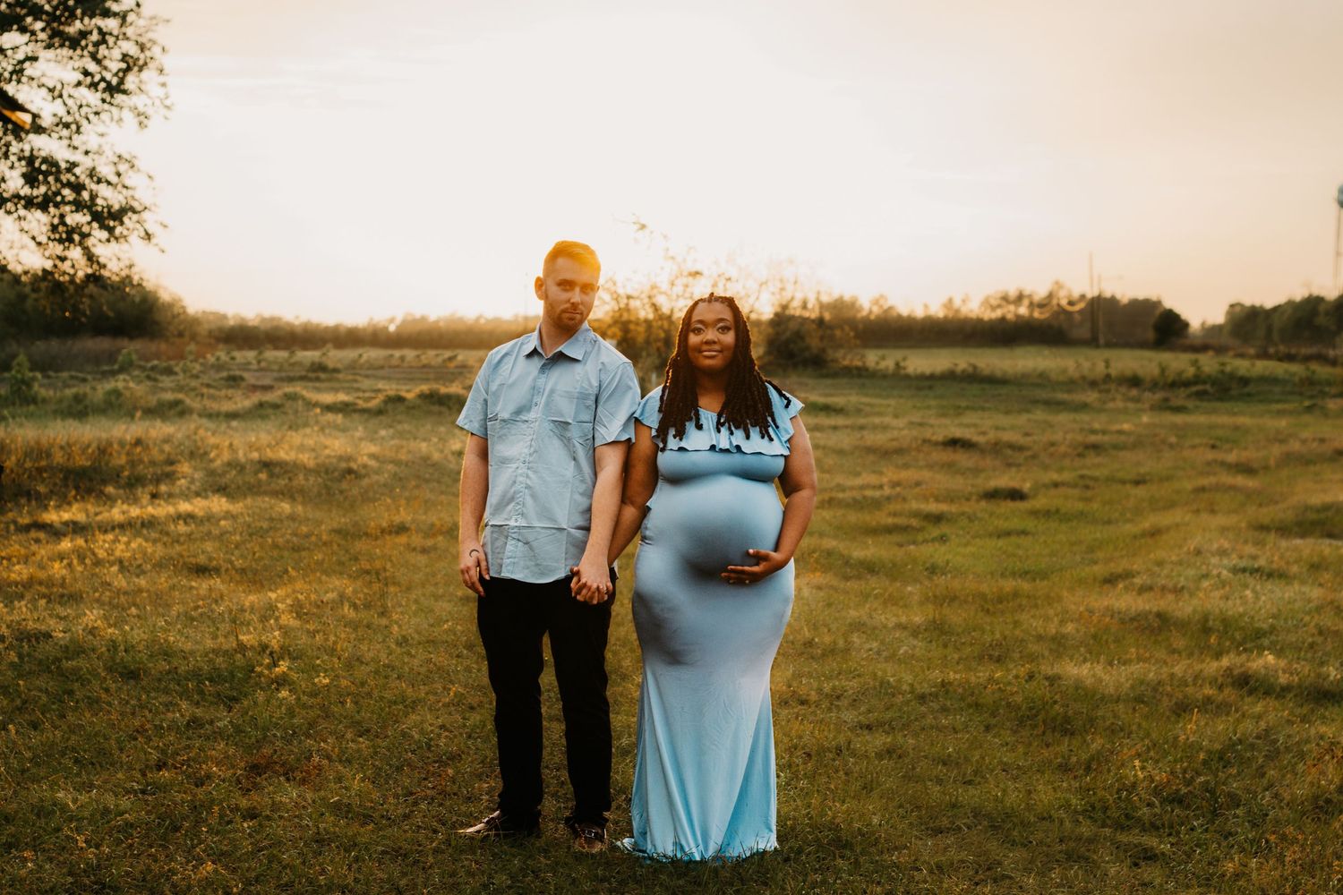 Smiling interracial couple holds hands in sunlit field, pregnant woman in blue maternity gown at sunset.