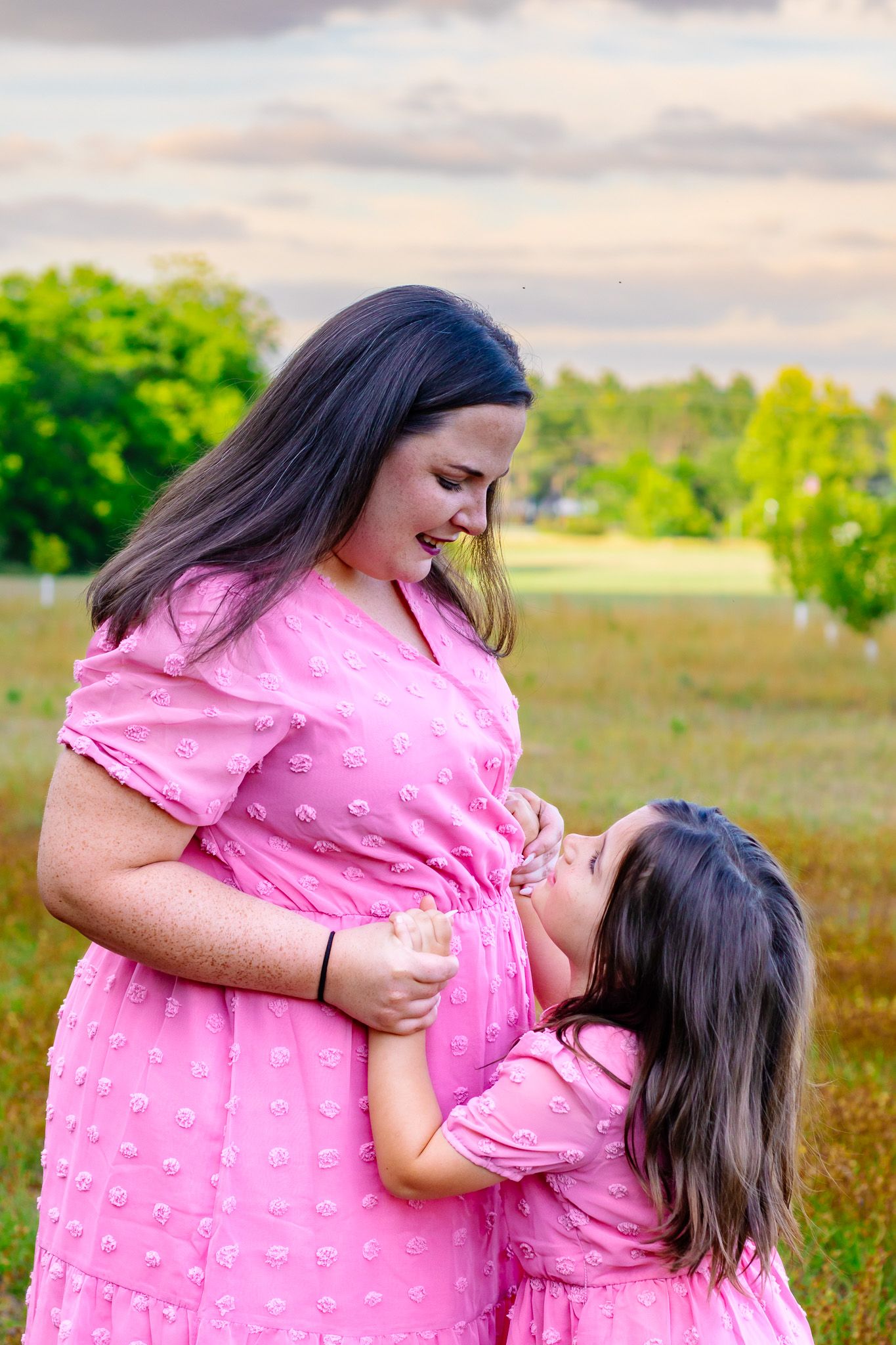 Pregnant woman and daughter in matching pink dresses sharing a tender moment outdoors in a green field.