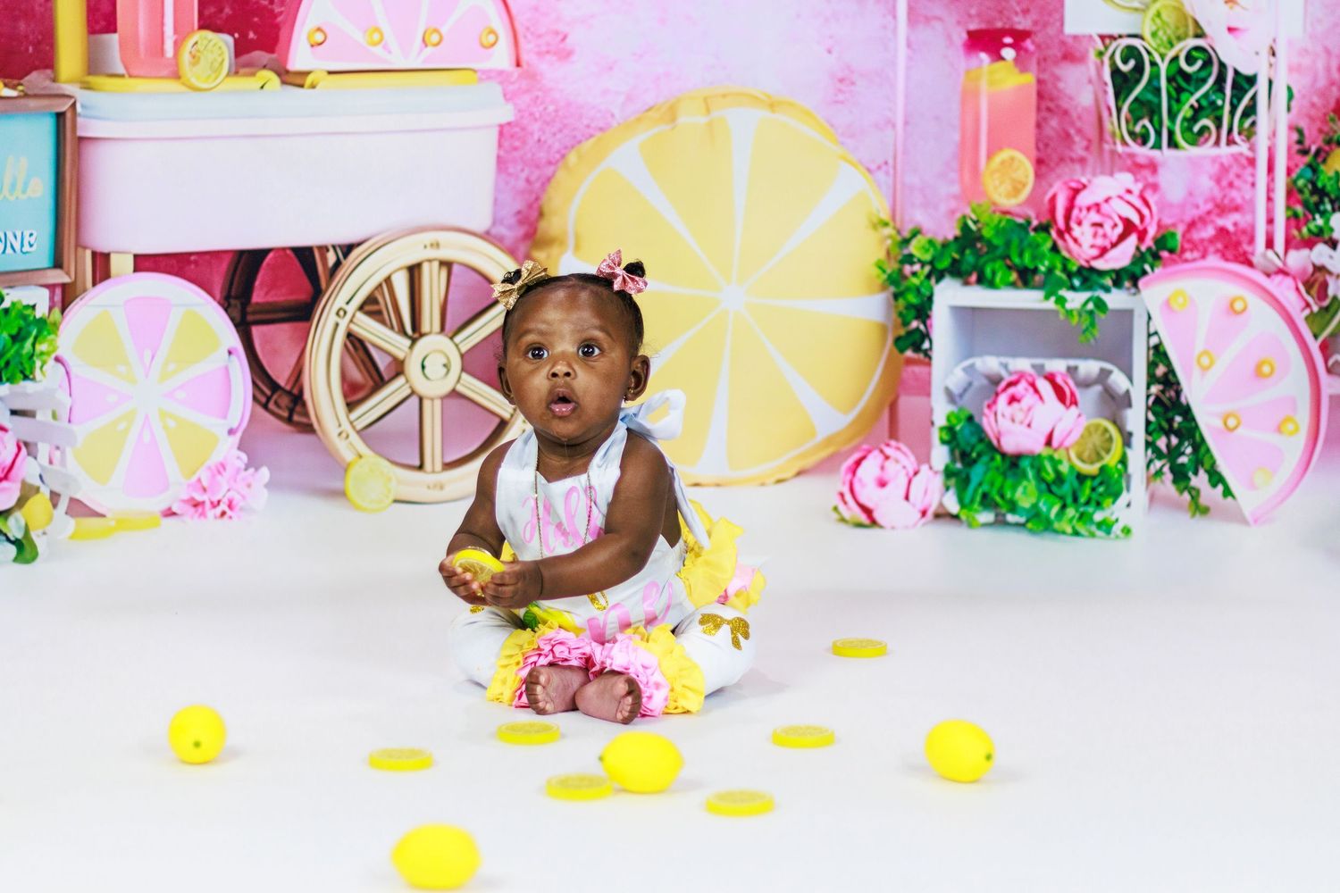 Baby girl in a pink and yellow dress sitting on white floor with lemon decorations and a colorful backdrop.