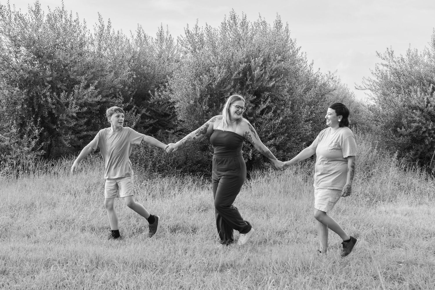 Black and white photo of three people holding hands and walking joyfully through a grassy field with trees in background.