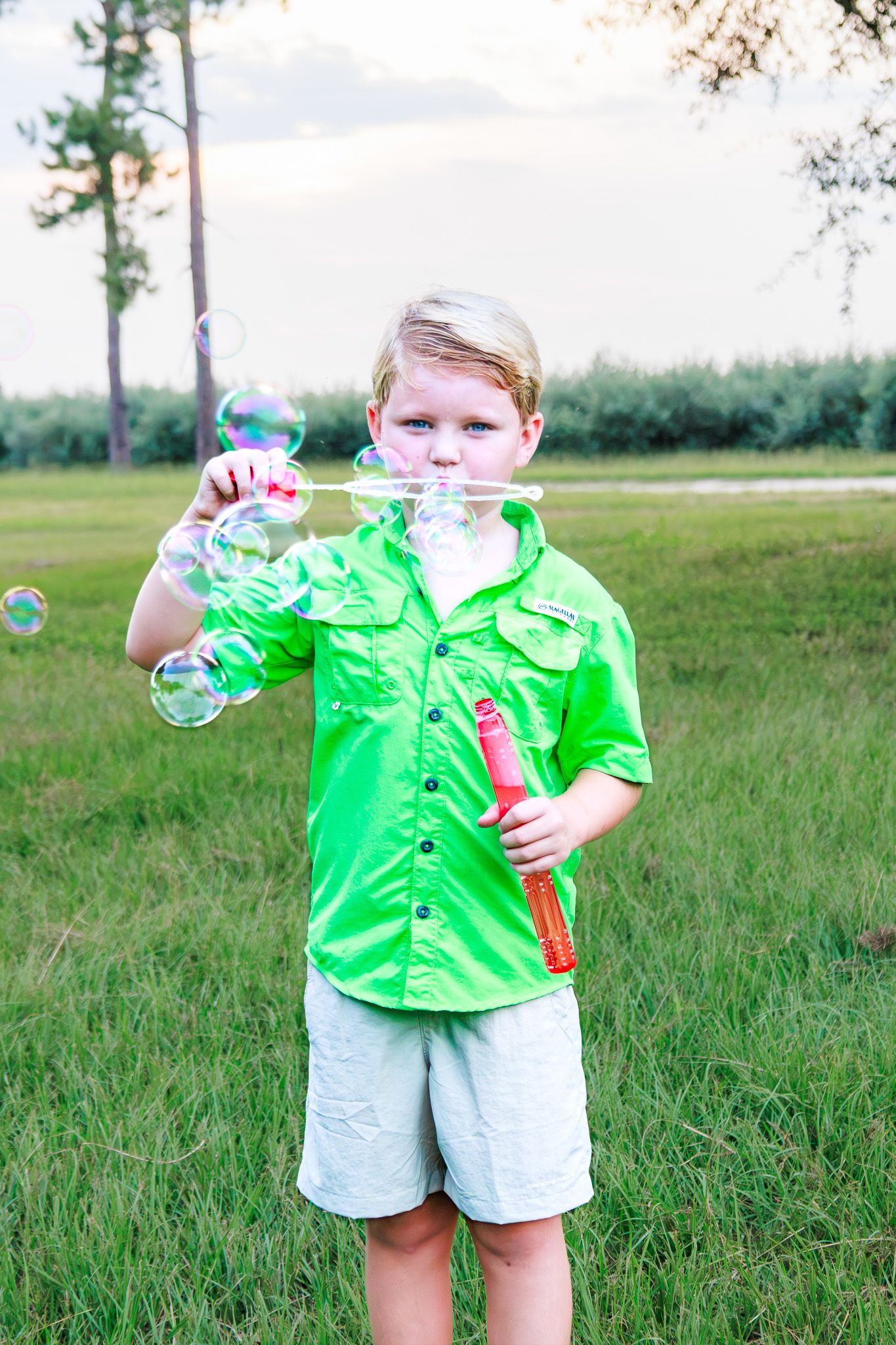 A young boy in a bright green shirt blows bubbles outdoors in a grassy field on a cloudy day.