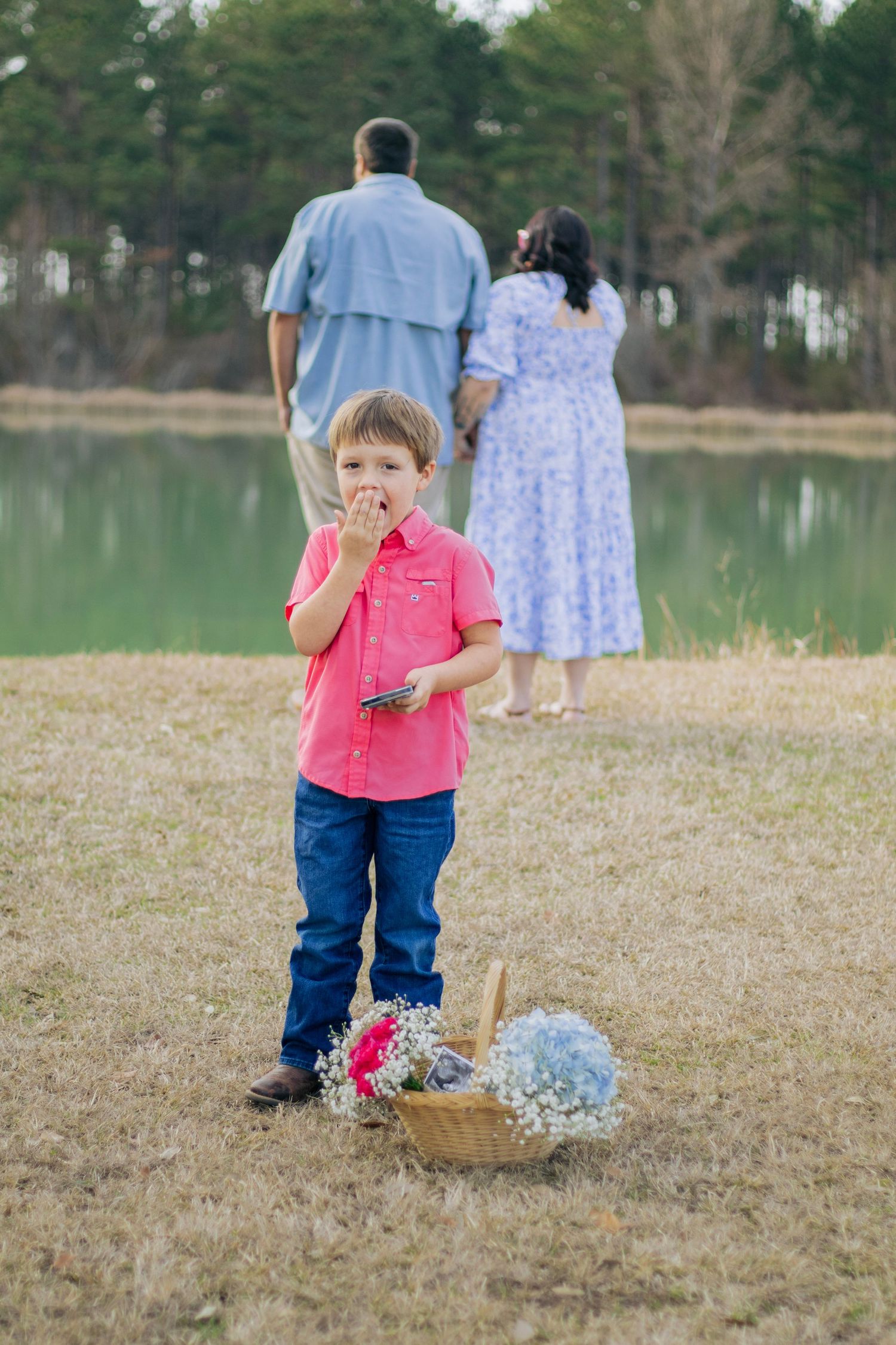 Young boy in pink shirt eating candy near basket of flowers by a pond.