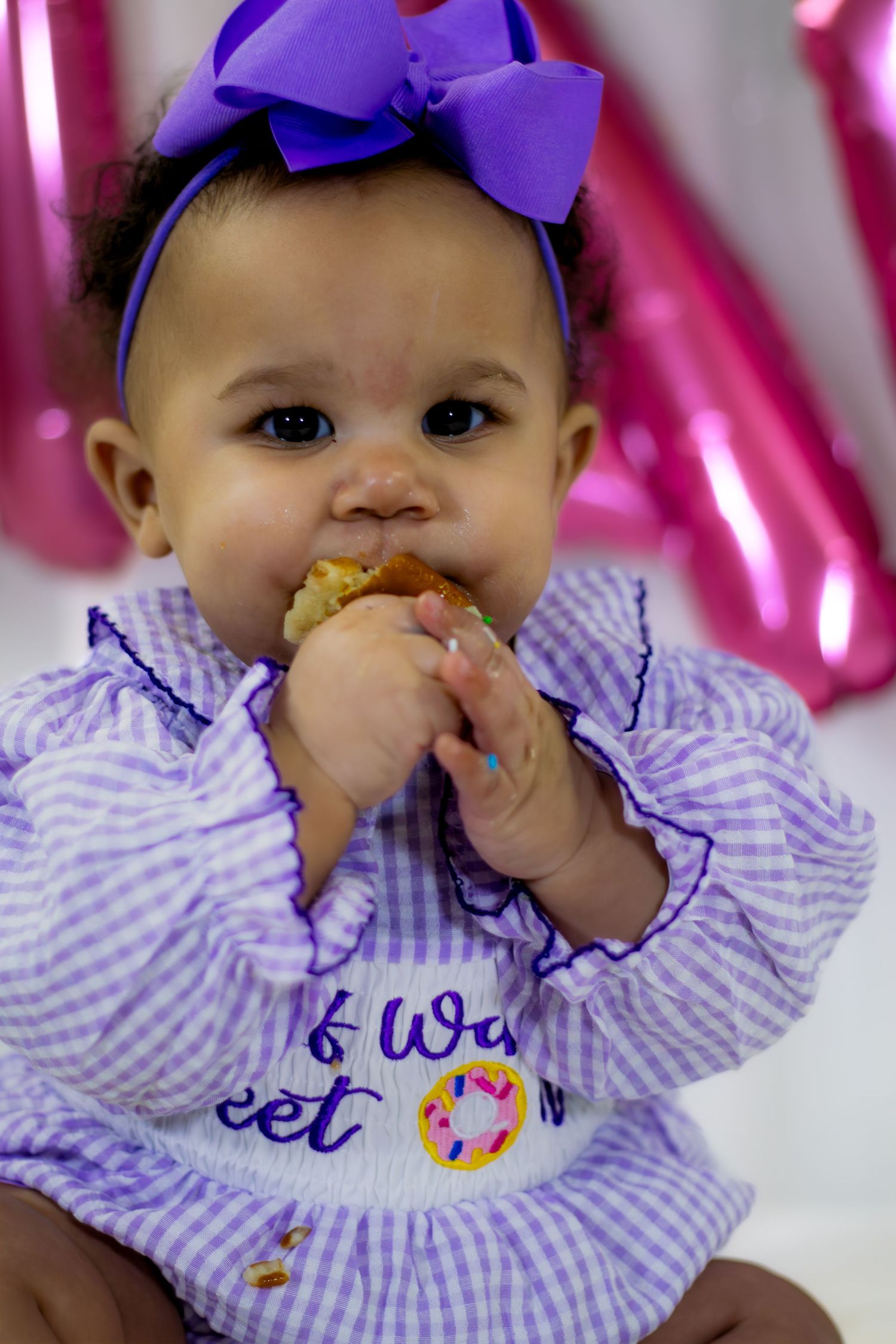 Baby girl in purple gingham dress with bow headband eating cake at her half birthday celebration.