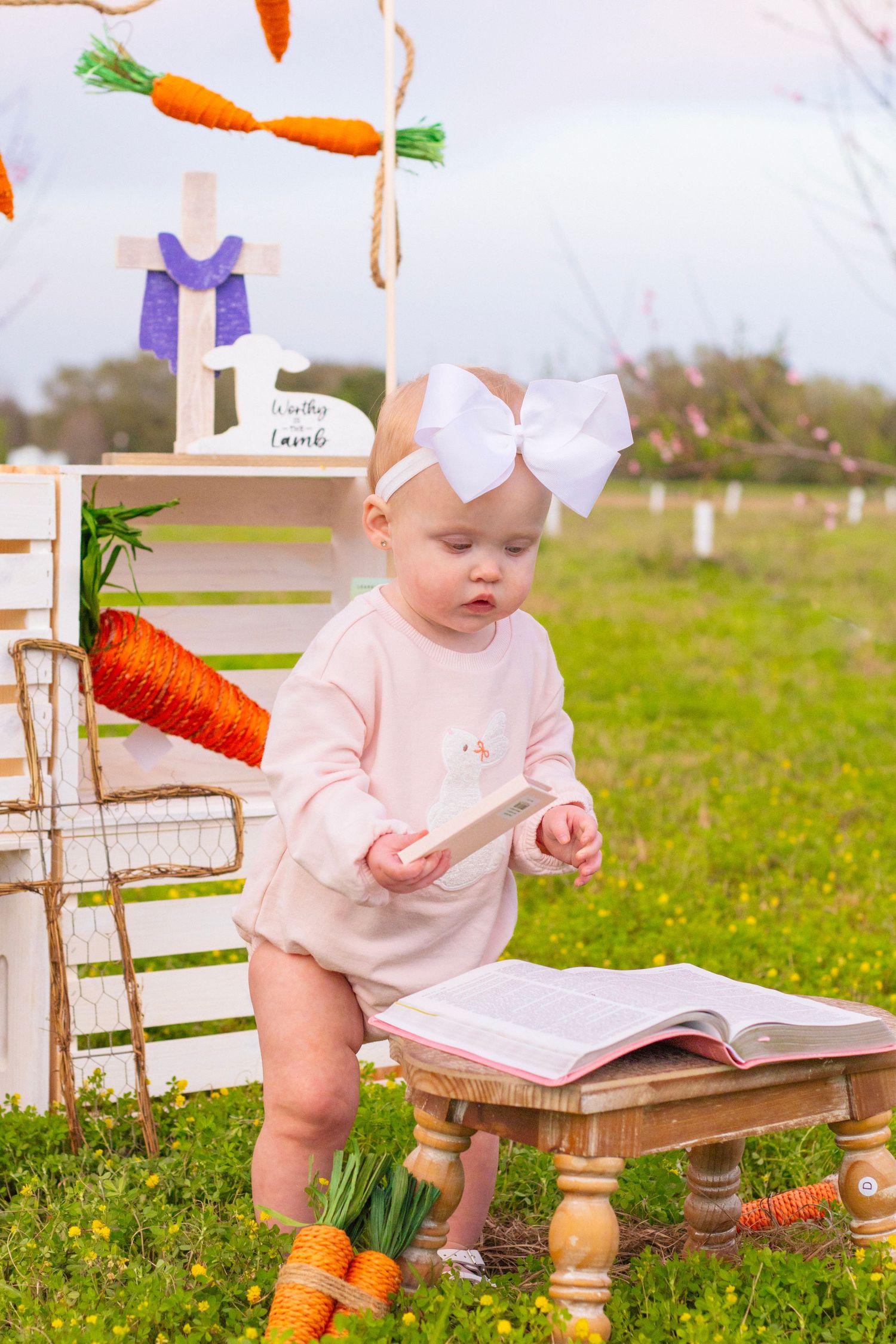 Baby with white bow headband reads open book at small table in Easter garden setting.