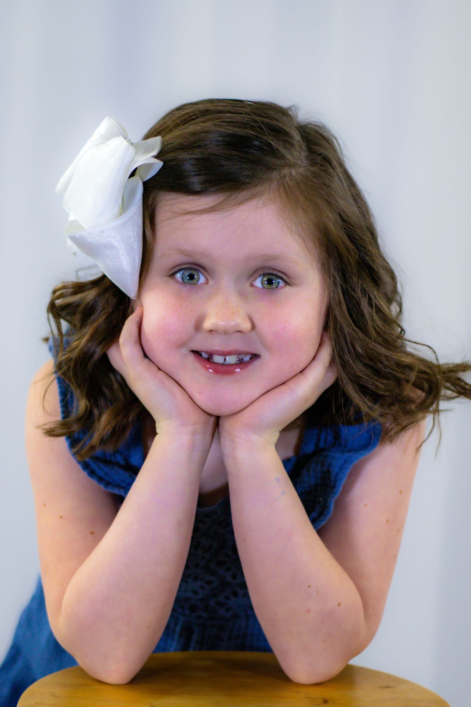 Adorable girl in blue dress resting chin on hands with big smile and white bow.